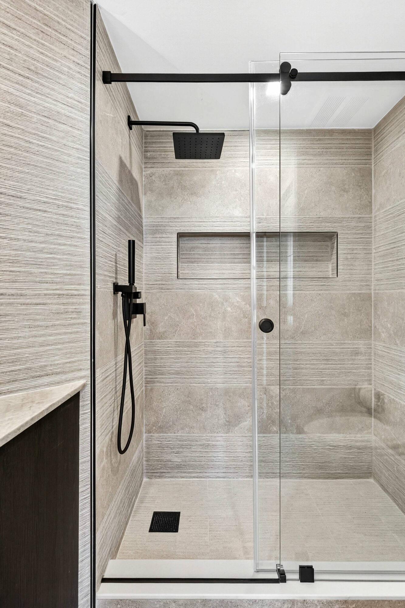 Shower area with black overhead rain shower, handheld shower, and built-in wall niches, enclosed by a glass door.