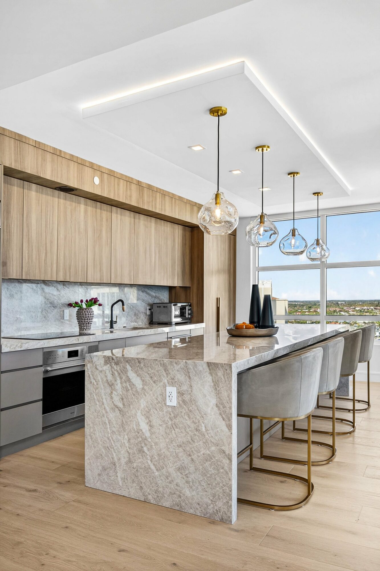 Modern kitchen with marble island, gray cabinets, wooden upper cabinets, and pendant lights, overlooking a city view.