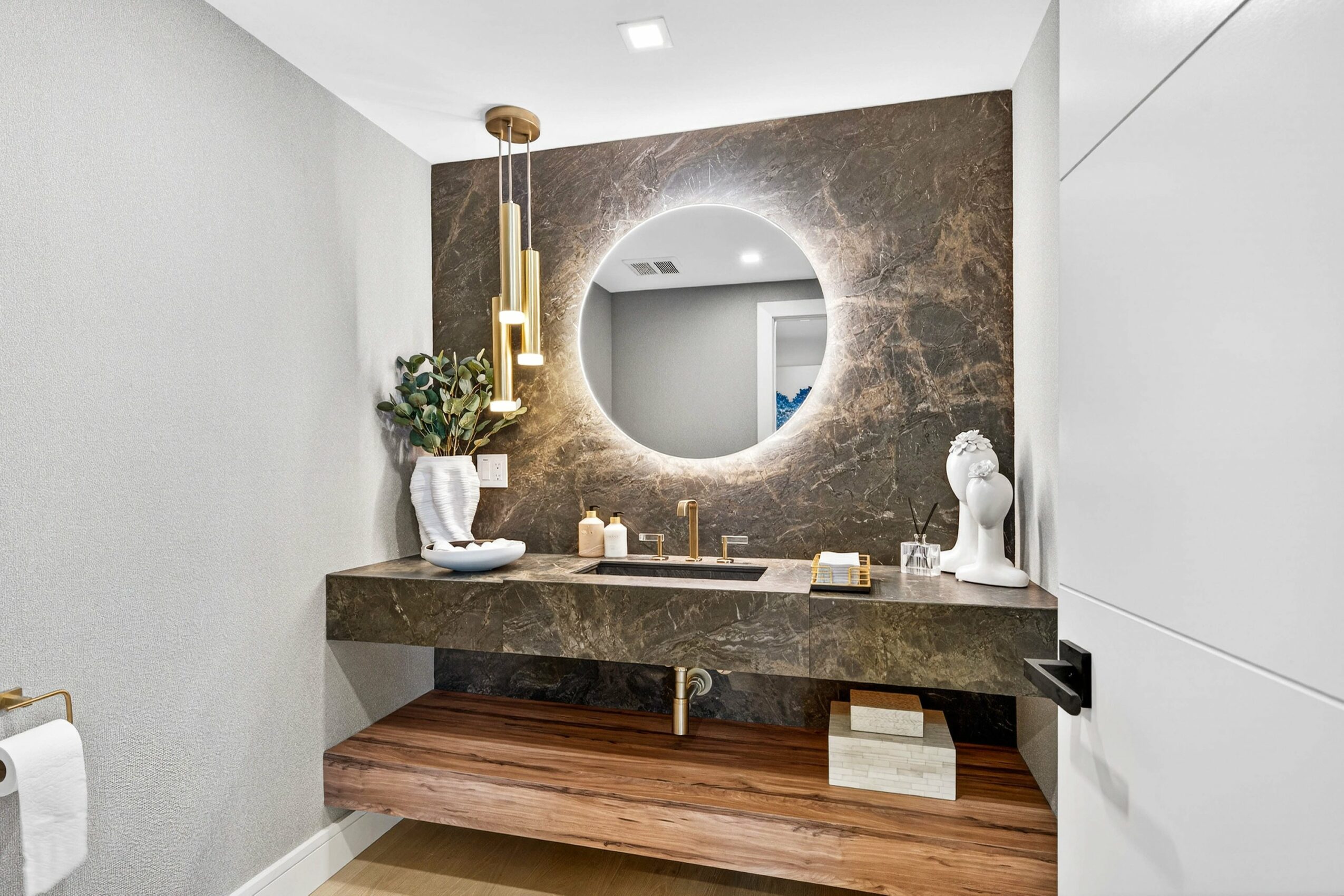 Bathroom vanity with round mirror, candles, and decorative sculptures, against a textured wall, with a wooden shelf below.