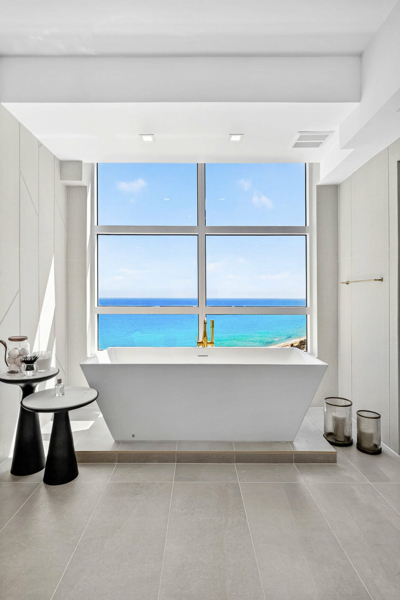 Bathroom with a bathtub facing large window showing ocean view, with two black stools and decorative items nearby.