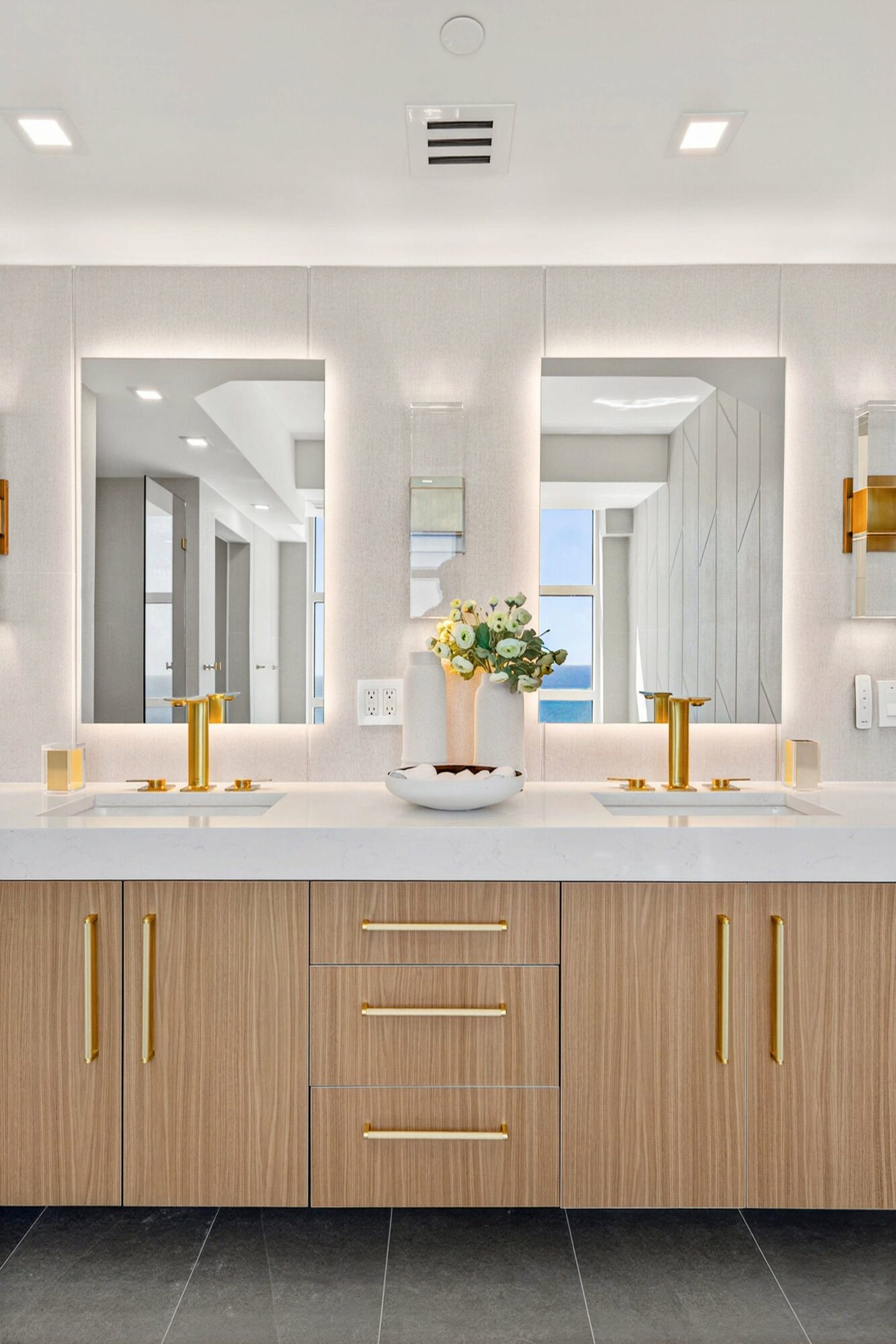 Bathroom vanity with two sinks, gold fixtures, a flower arrangement, and large mirrors, with a bright, modern background.