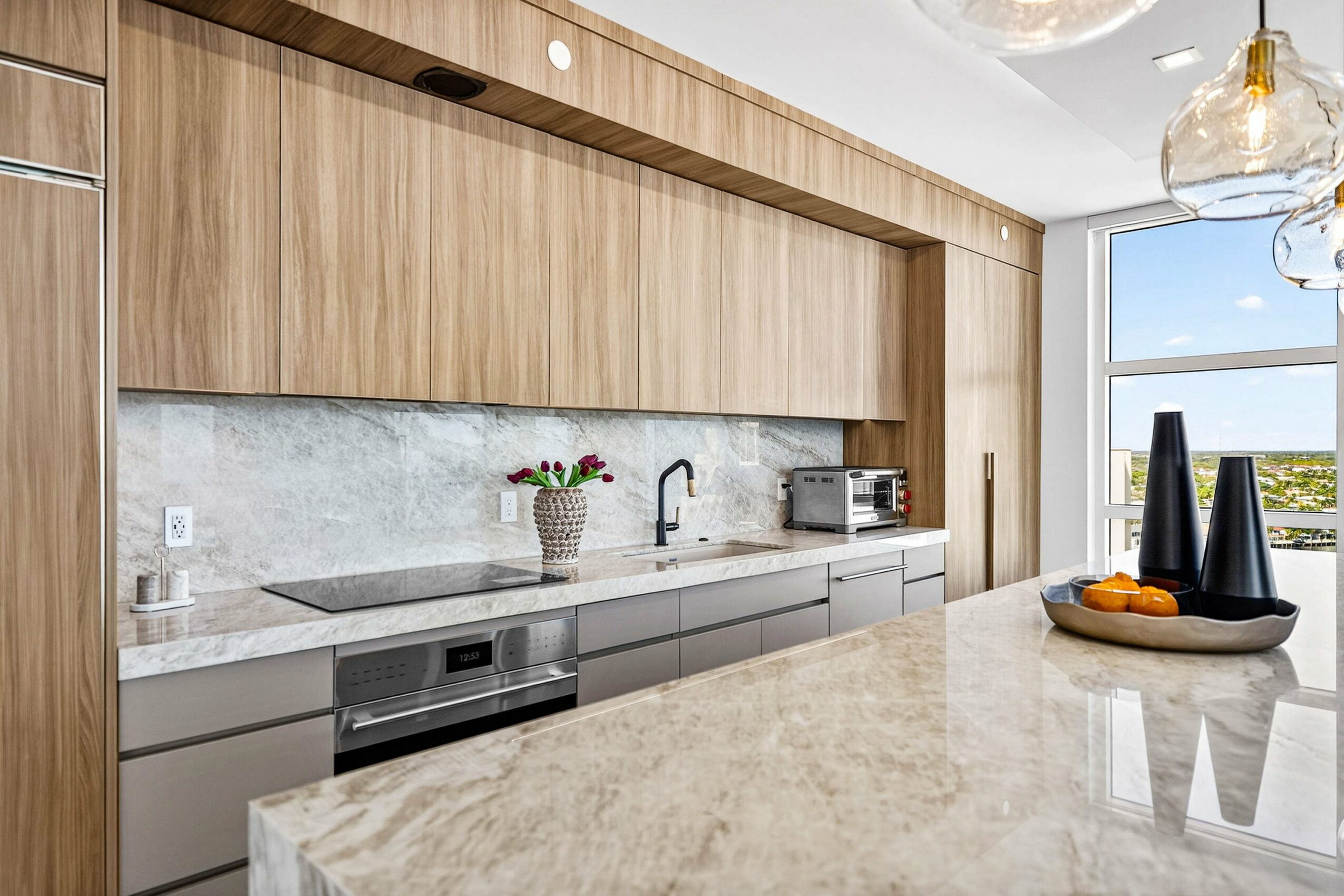 Modern kitchen with wooden cabinets, gray lower cabinets, marble countertop, and a window with a view outside.