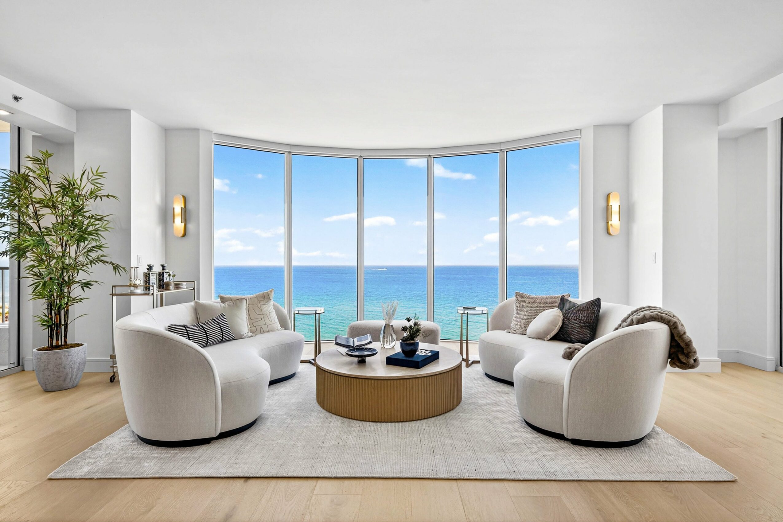 Living room with large windows showing ocean view, two white curved sofas, a round coffee table, and a potted plant.