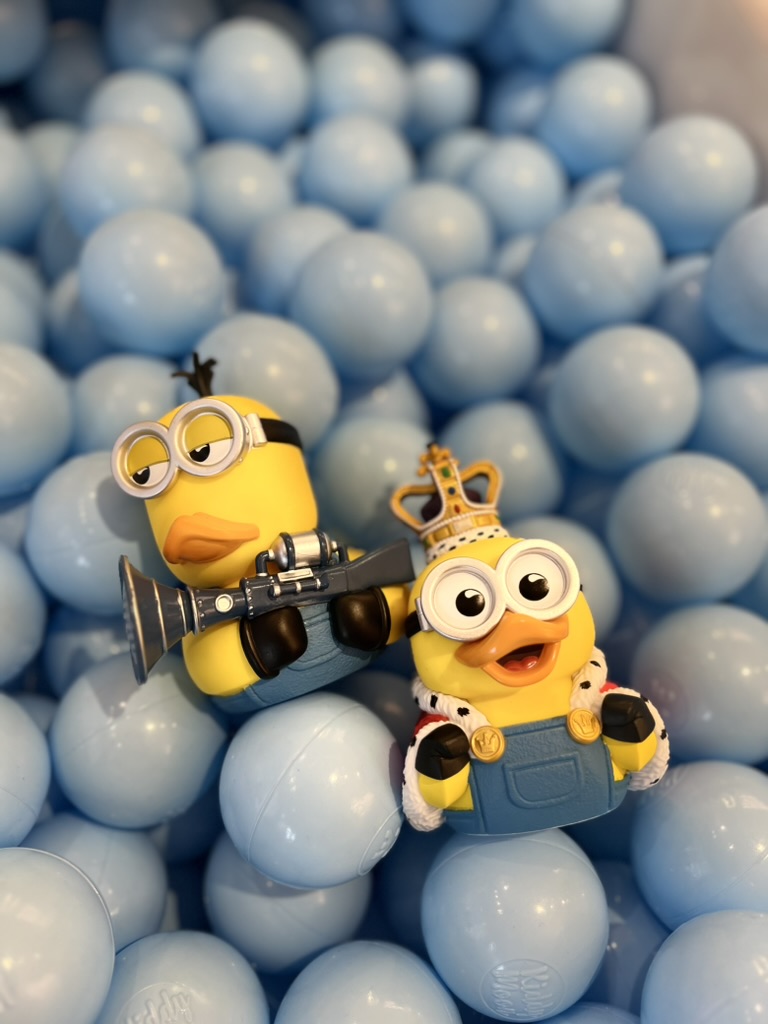 Two Minion toys among light blue balls, one with goggles and a camera, the other with a crown and overalls.