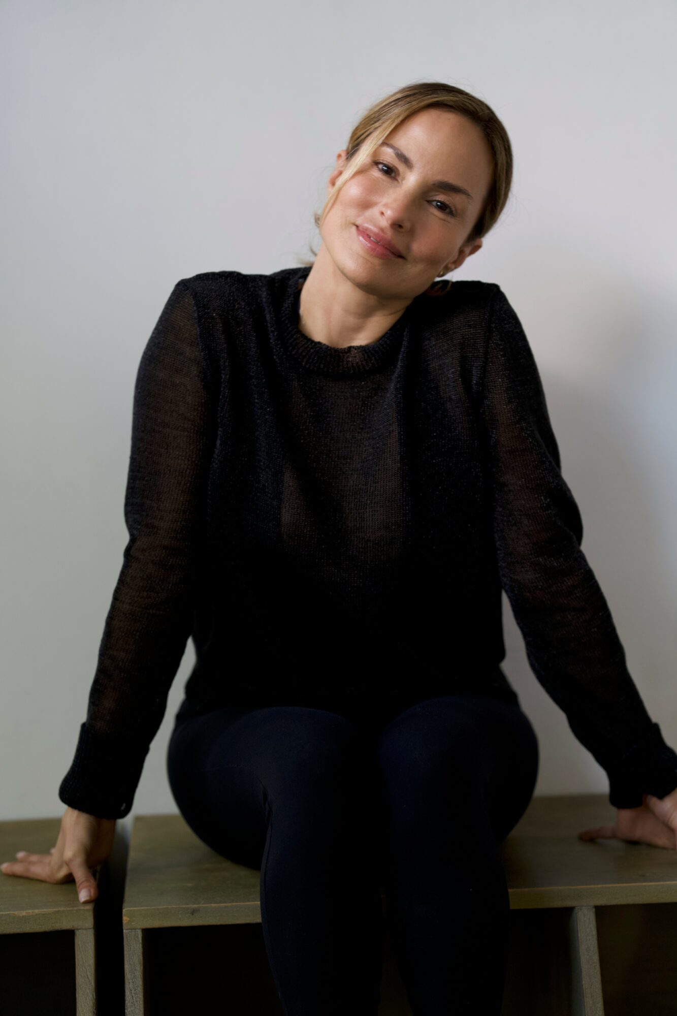 Young woman with light brown hair in a ponytail, wearing a black long-sleeve top, sitting on a wooden surface, smiling gently.