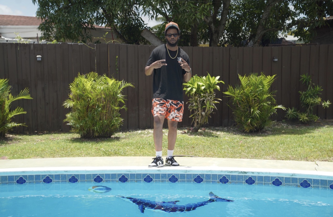 Man standing by a swimming pool with green bushes and a wooden fence in the background.