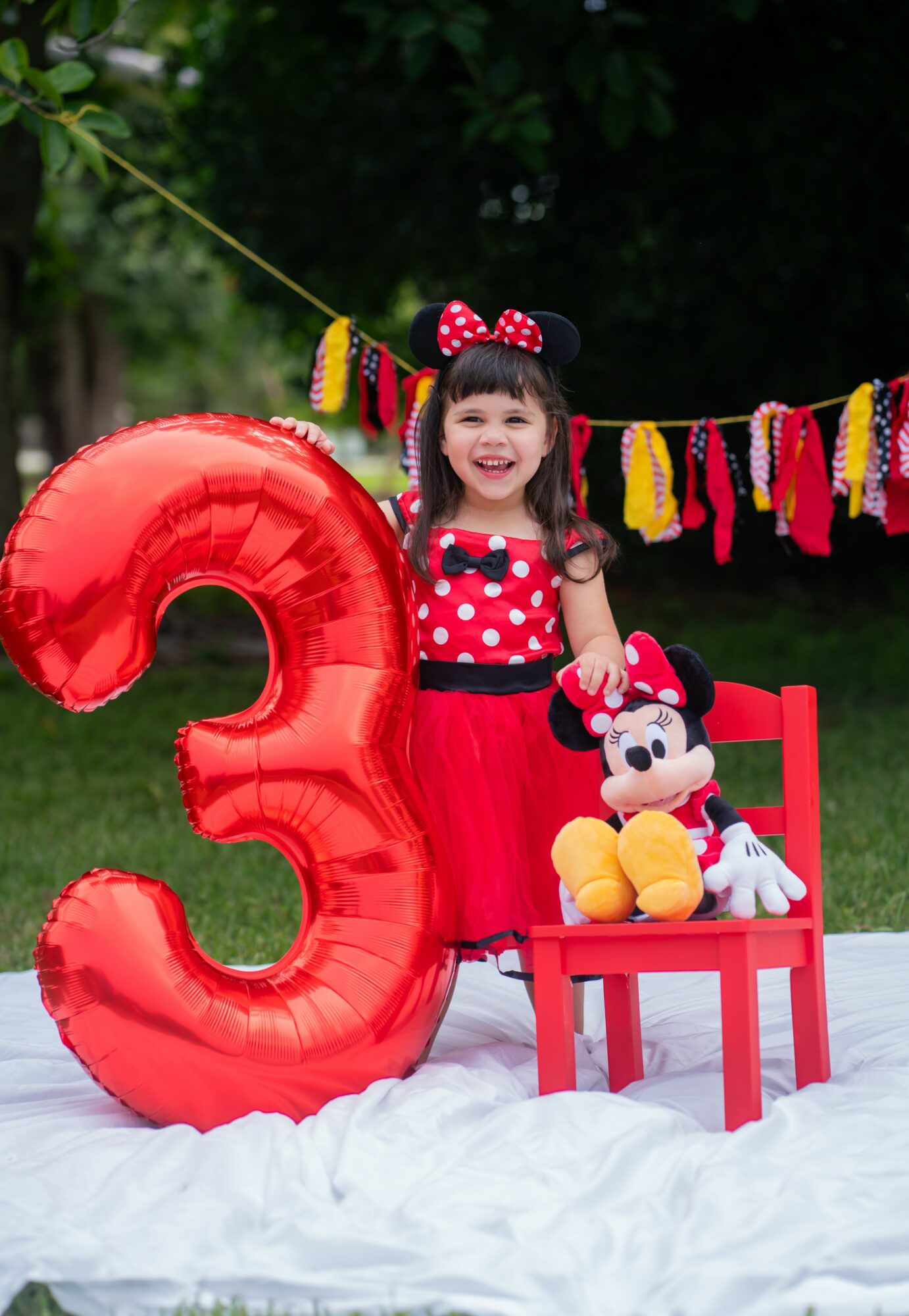 Young girl in Minnie Mouse dress with Minnie plush toy, sitting on a red chair outdoors, celebrating third birthday with a large red number three balloon.