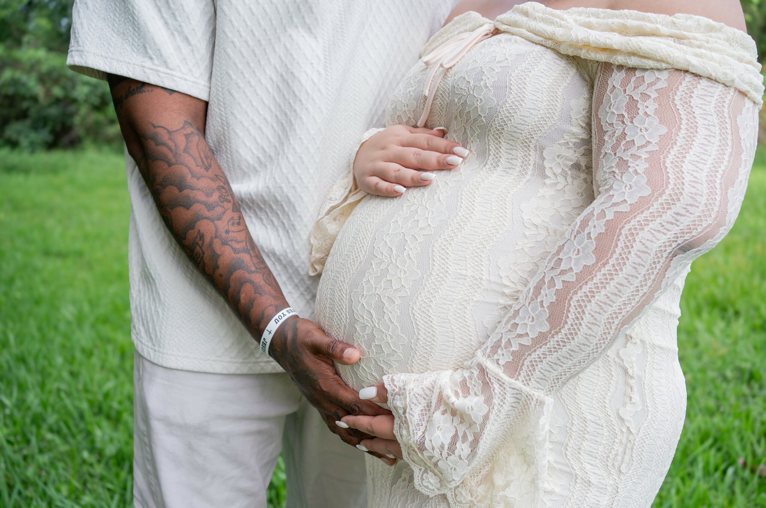 Close-up of a couple holding a pregnant woman's belly outdoors, with the man's hand on top of her belly, both wearing light-colored clothing.