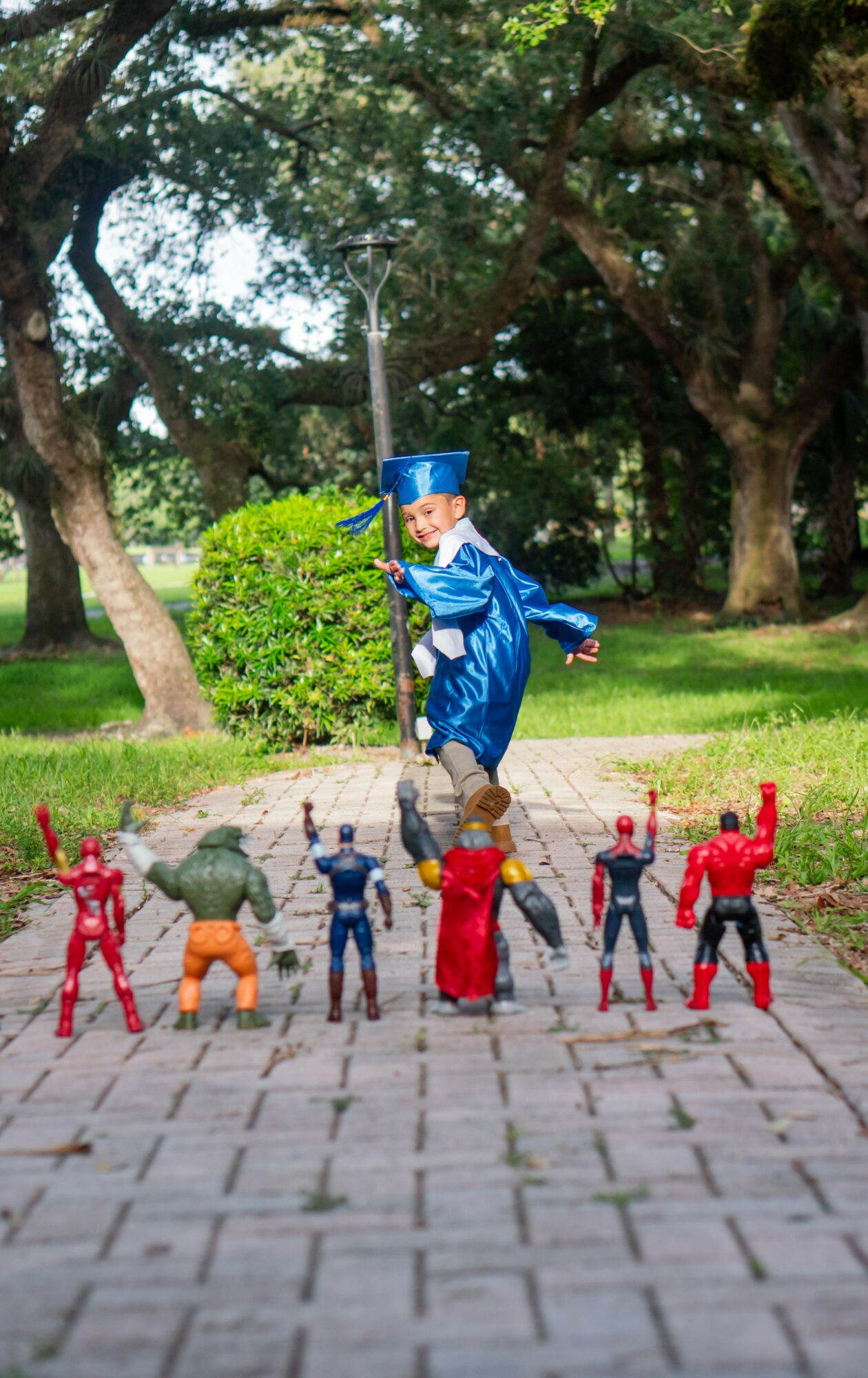 Child in a blue graduation gown and cap jumping over toy action figures on a paved path in a park, trees in background.