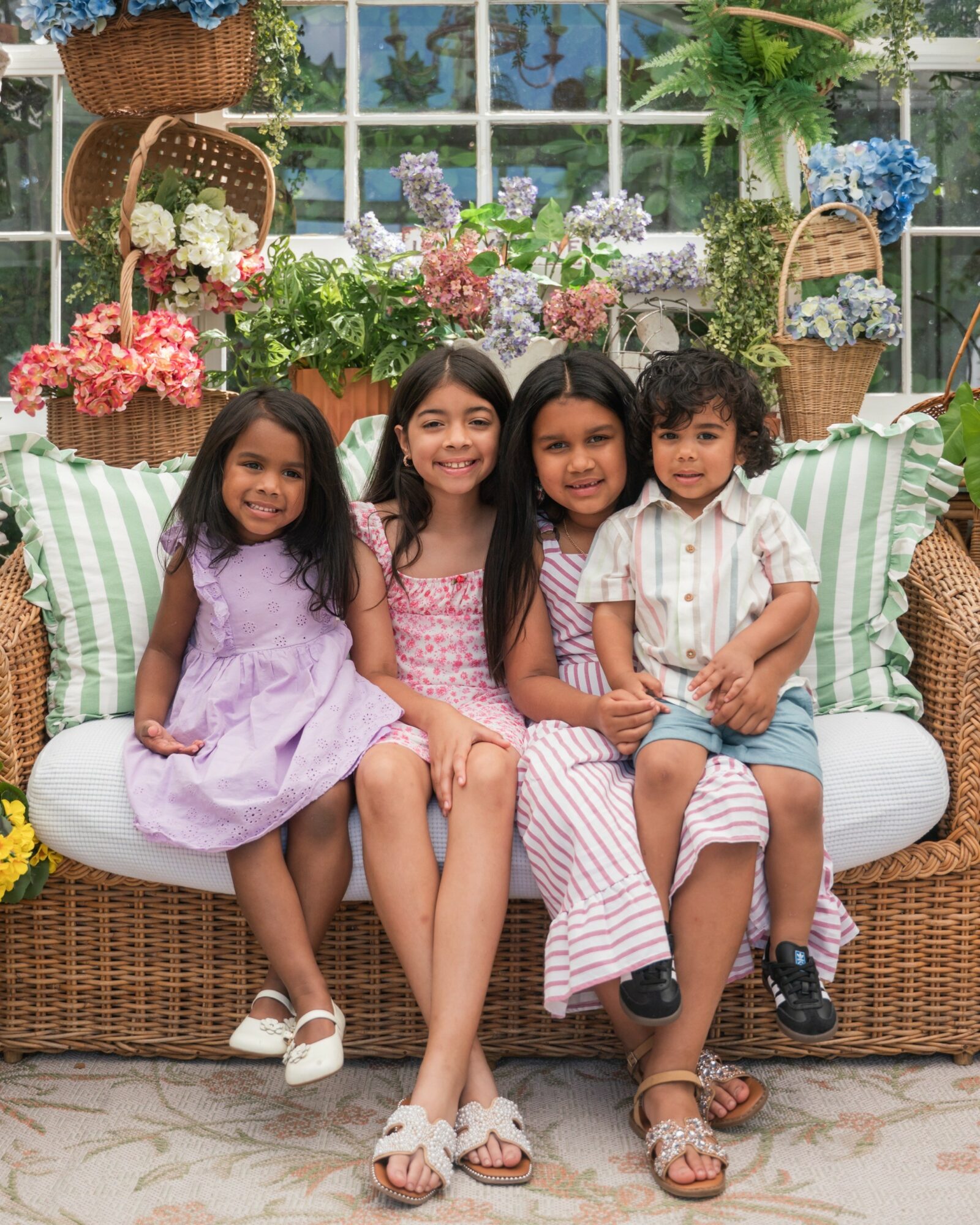 Four children sitting on a wicker sofa with floral decorations and greenery in the background.