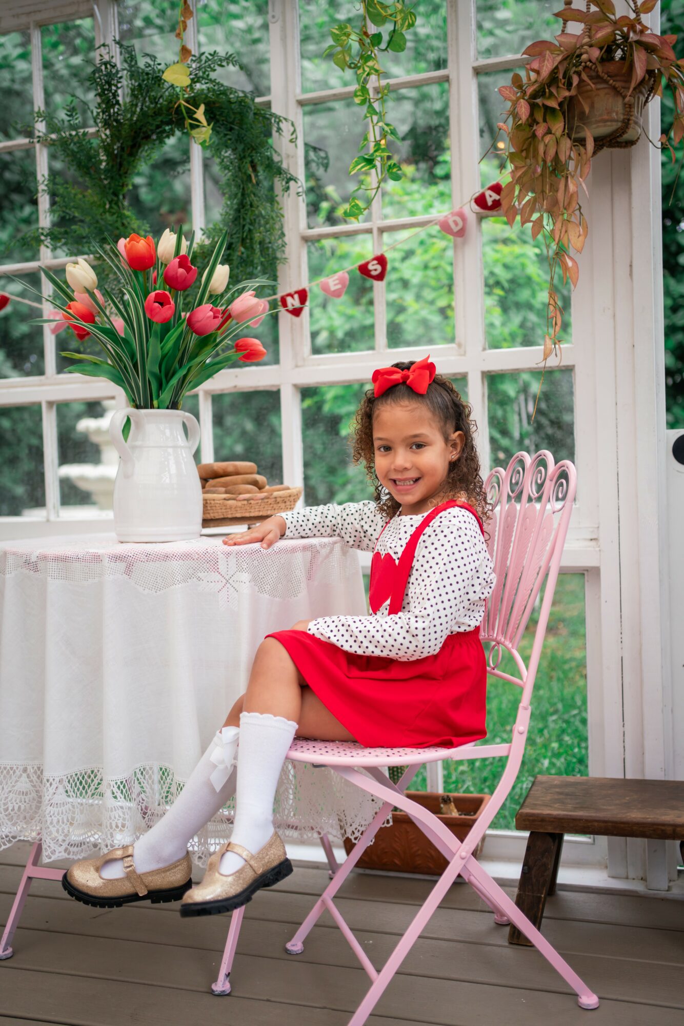 Young girl in red dress and white socks sitting on pink chair at decorated table with flowers, indoors with window and greenery.