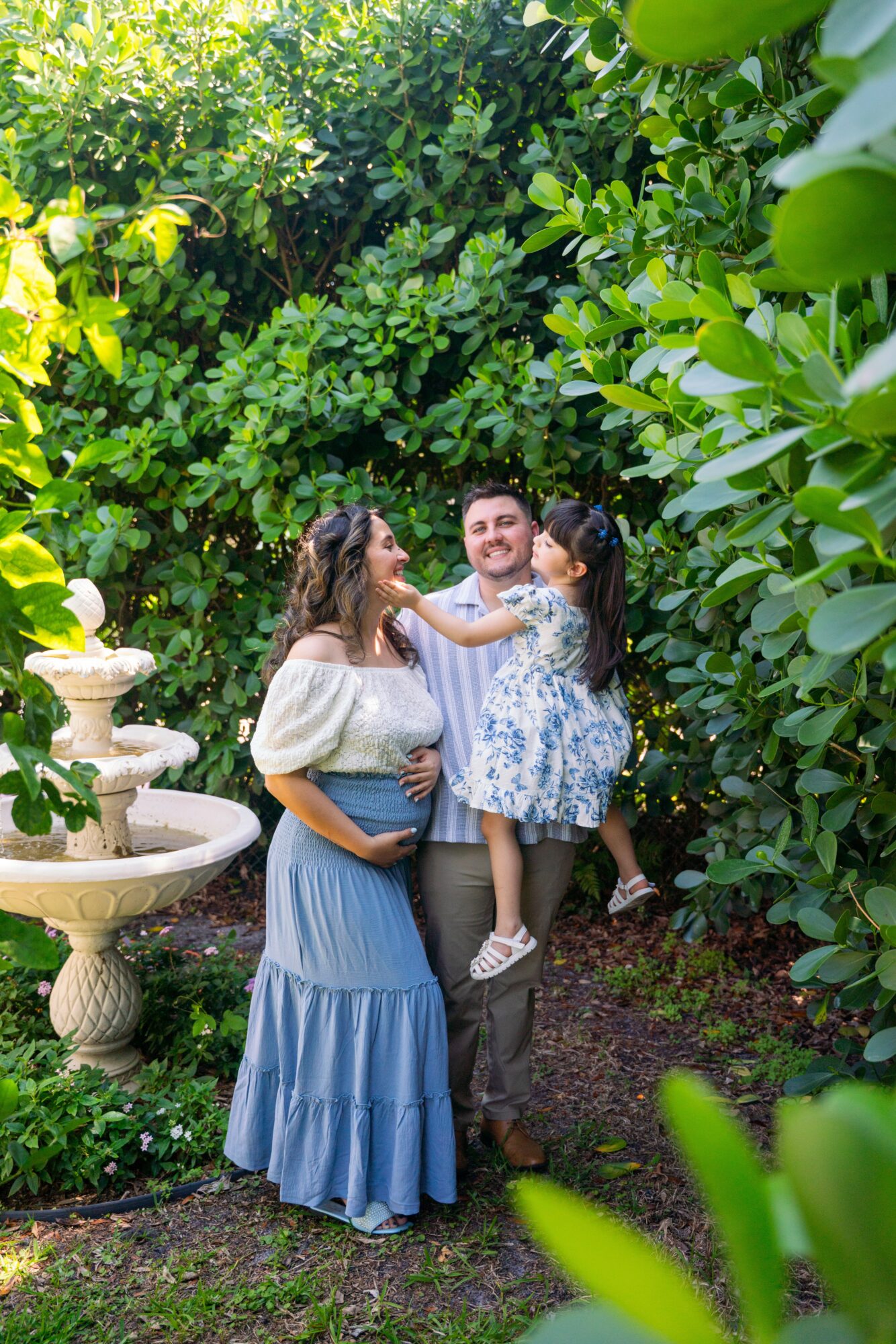 Family of four in a lush garden with greenery and a fountain, smiling and playing together.