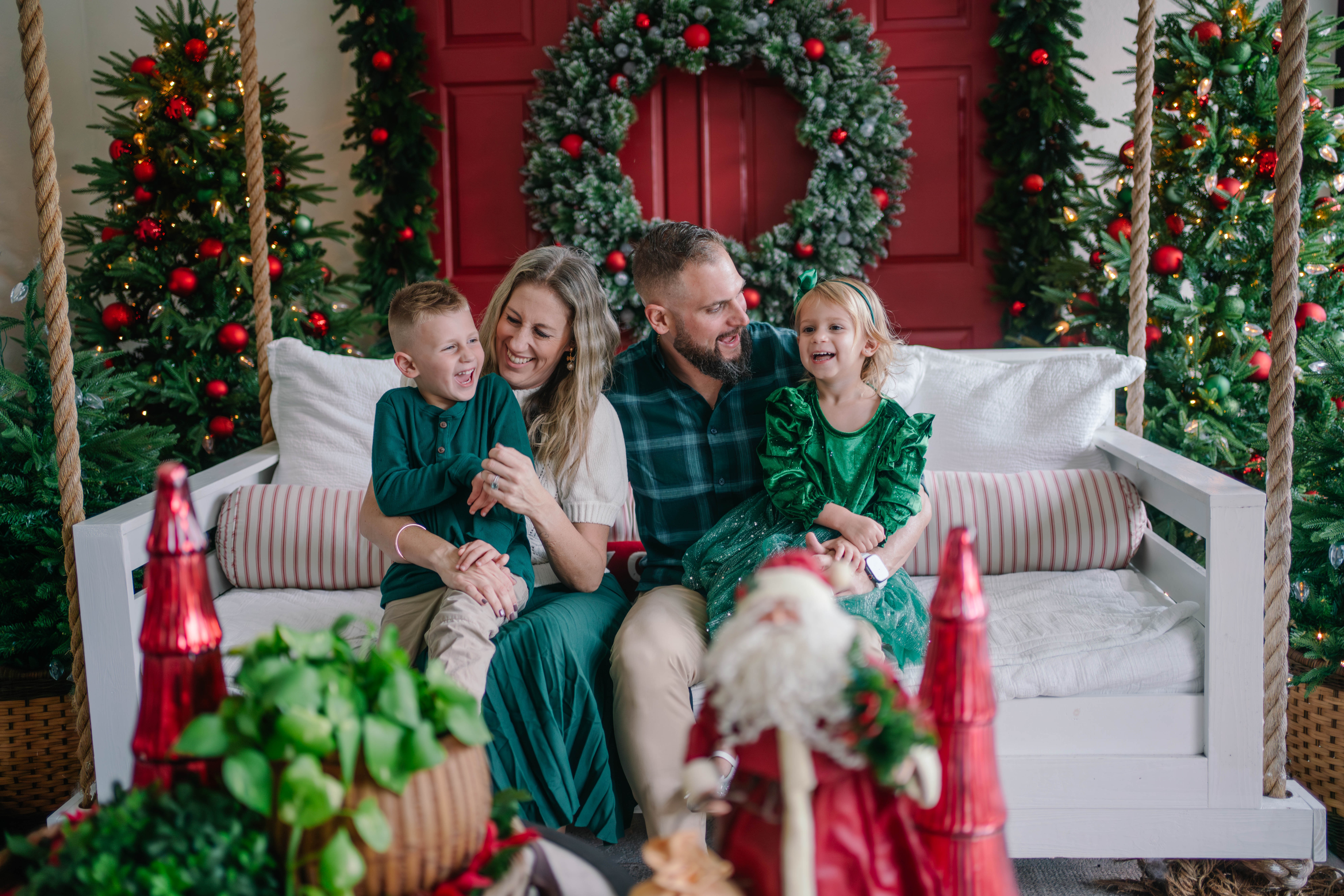 Family sitting on a decorated holiday sofa with Christmas trees and wreaths, smiling and enjoying the festive setting.