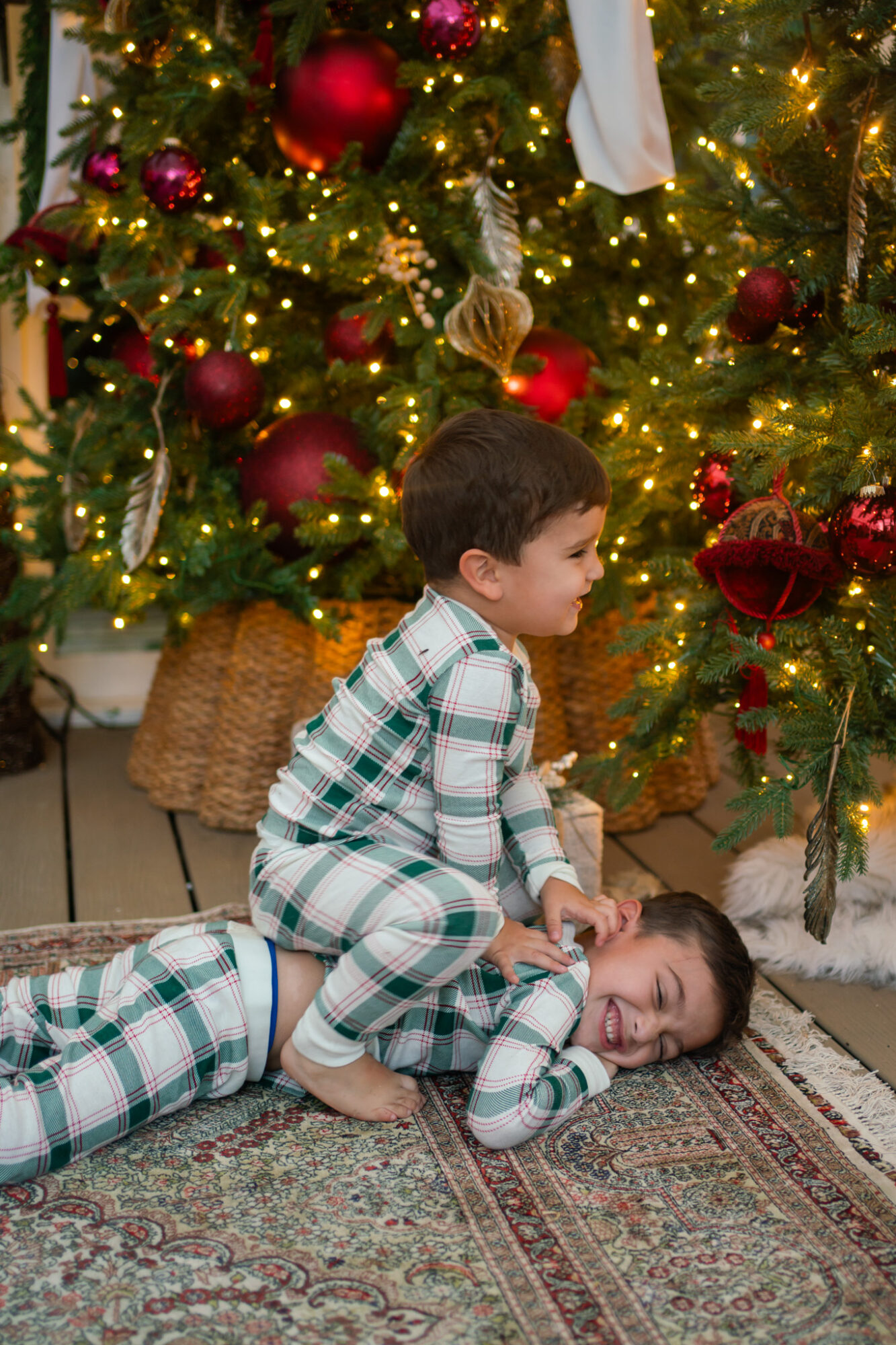 Two children playing on a decorated Christmas tree with ornaments and lights, one lying down and the other sitting on top.