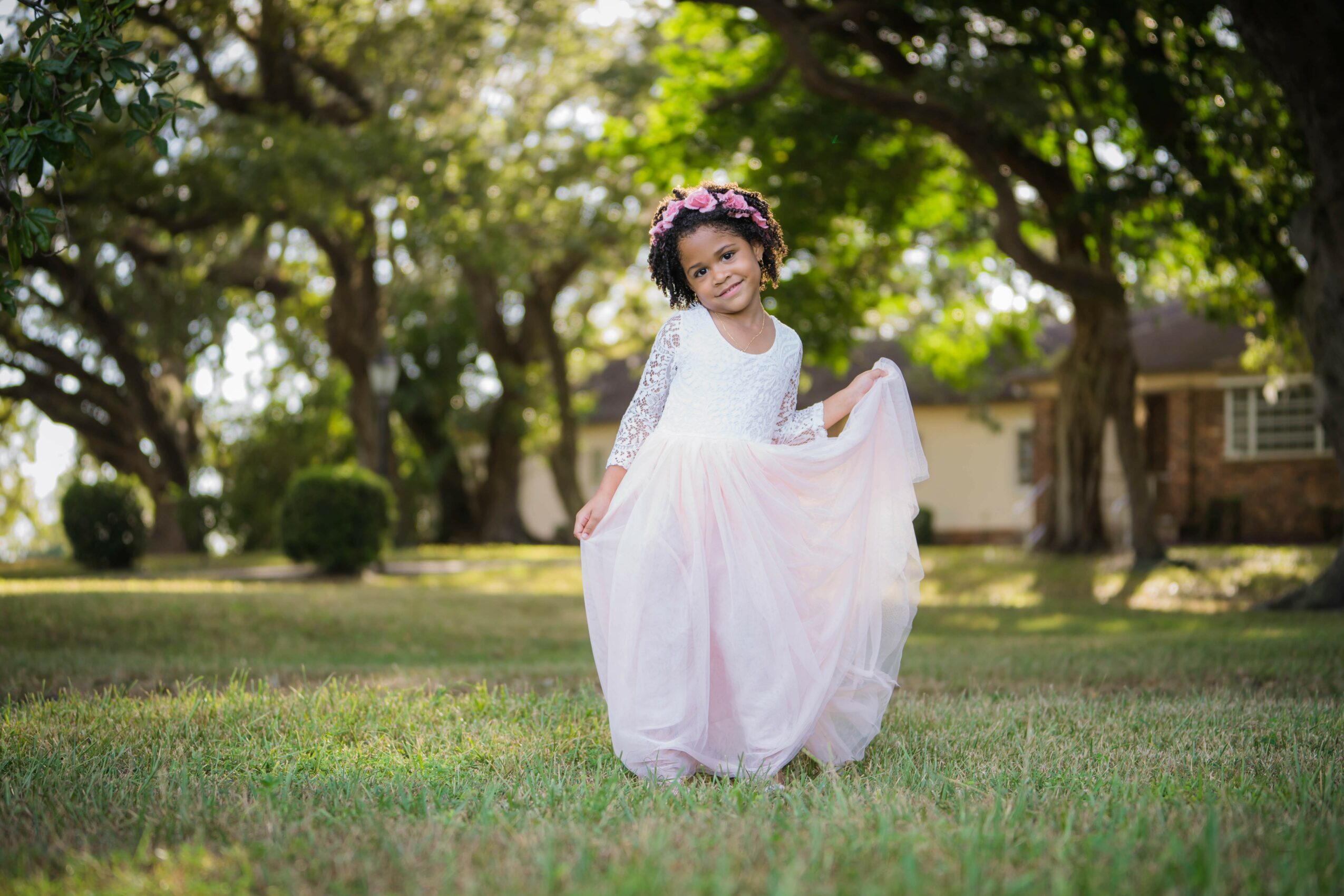Young girl in a pink dress and floral crown standing on grass in a park with trees and houses in background.