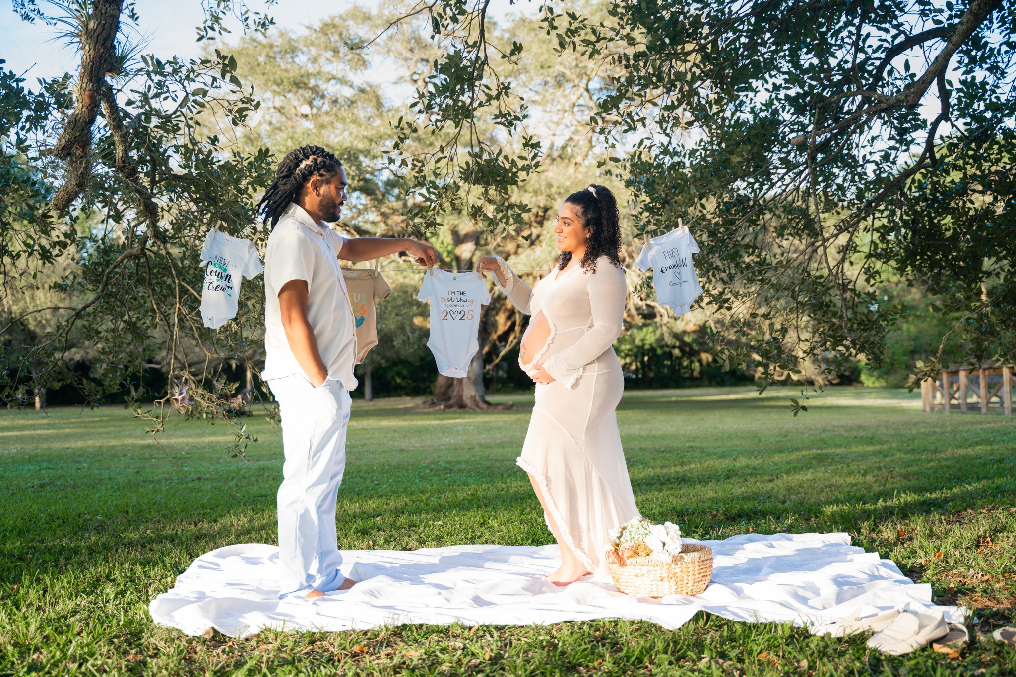A couple standing on a white cloth outdoors, holding hands, with trees and decorations in the background.