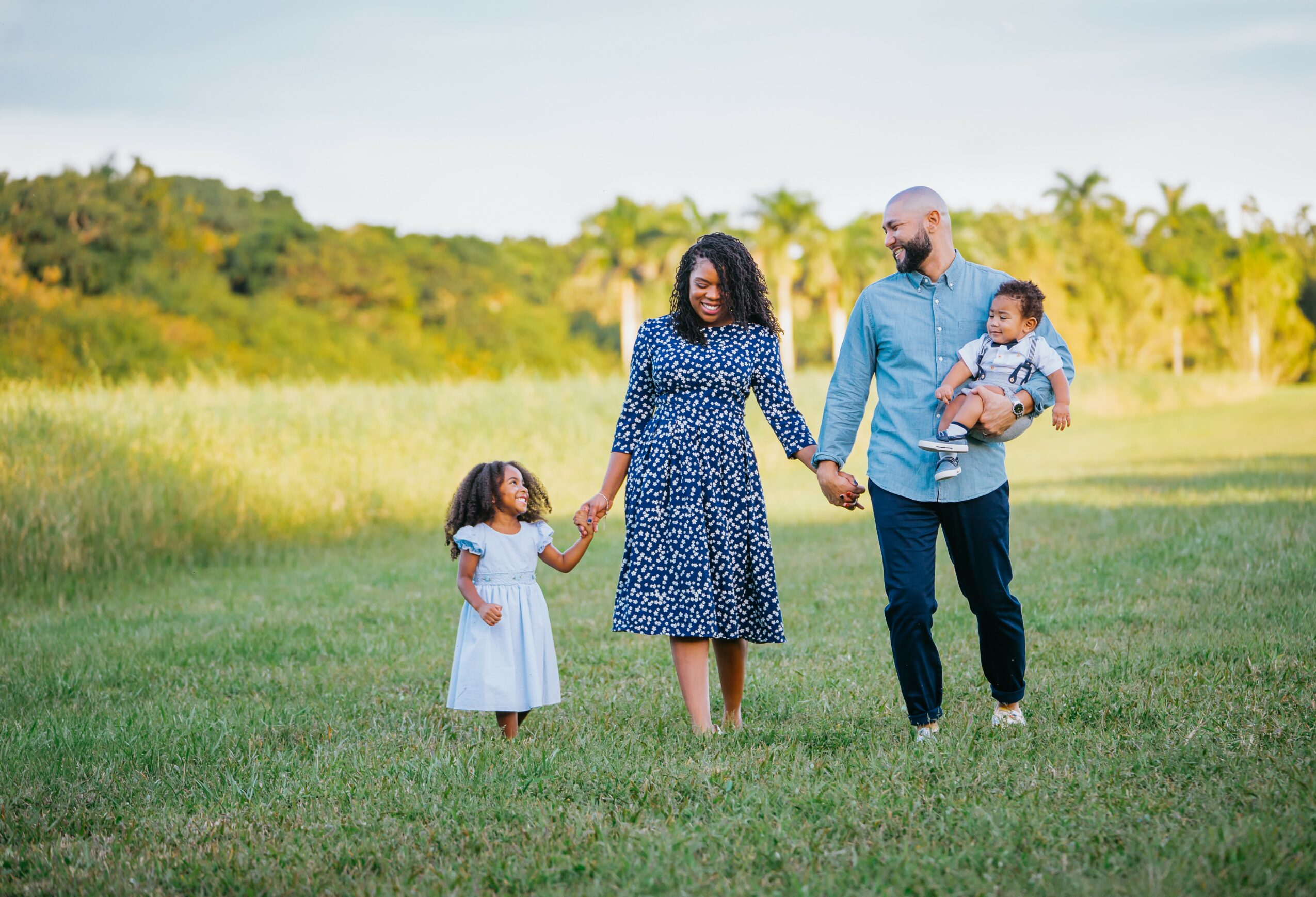 Family of four walking in a grassy field holding hands, with trees and blue sky in the background.