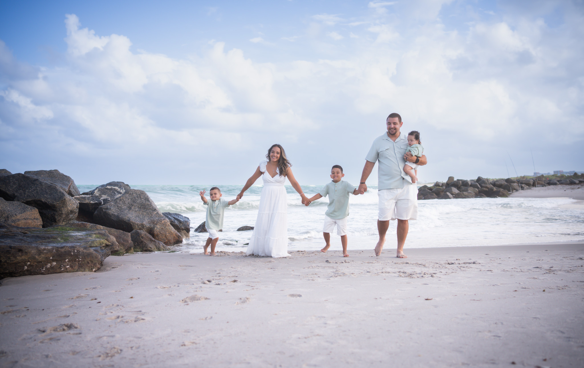 Family of five holding hands walking on beach with rocks and ocean in background, sky with clouds, sunny day.