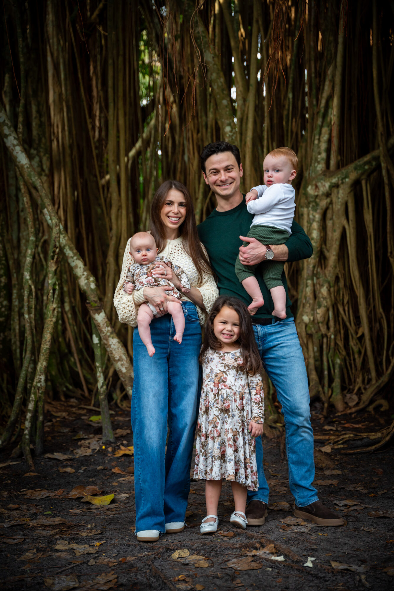 Family of five standing outdoors in front of large tree roots, smiling, with two children and a baby, in casual clothing.