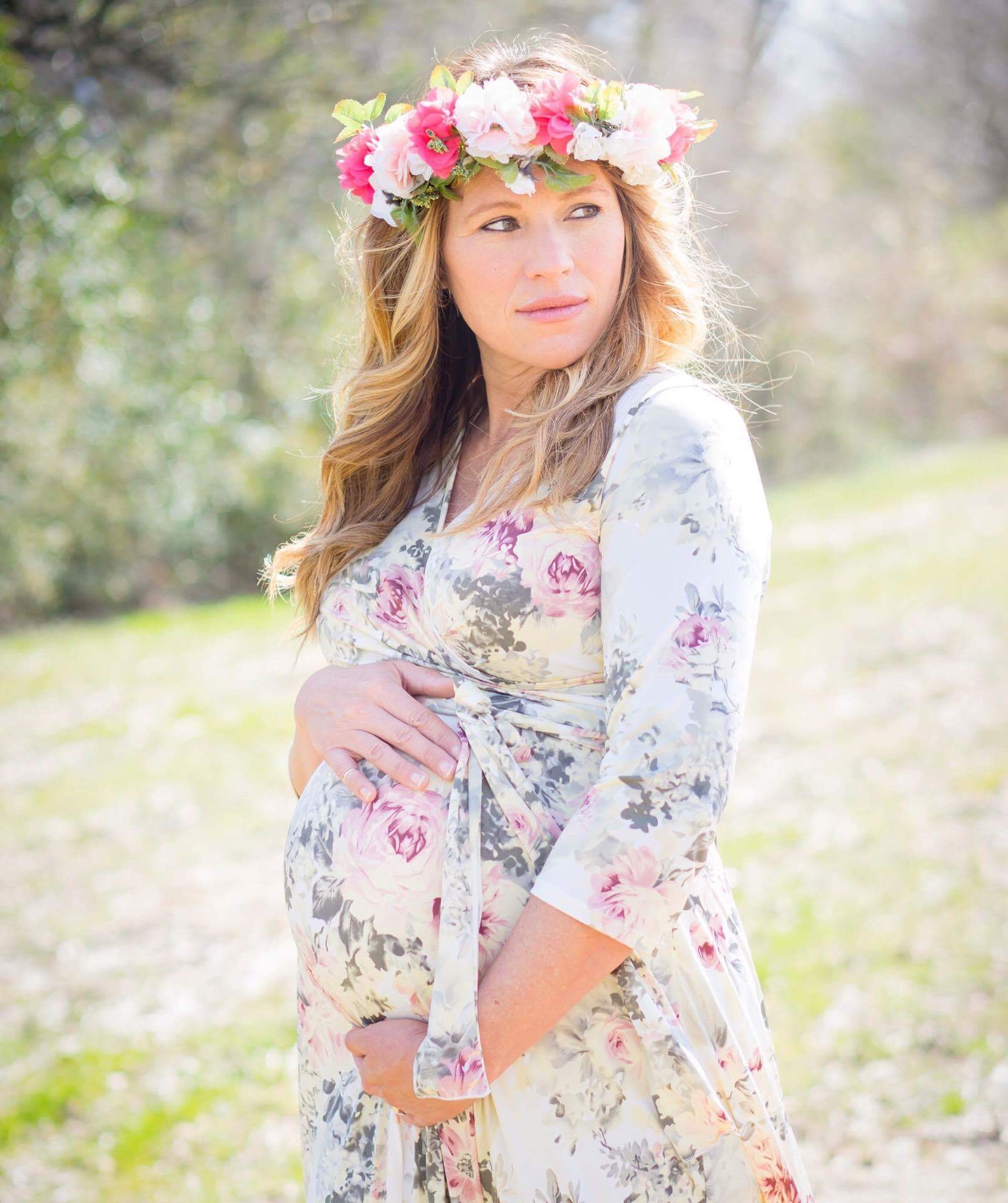 Pregnant woman outdoors wearing a floral dress and a flower crown, looking to the side, with a blurred natural background.