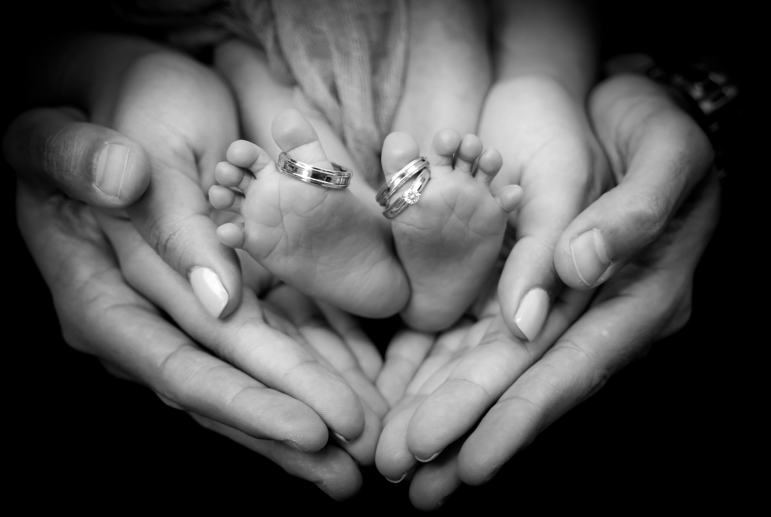 Adult hands gently hold a baby's feet with rings on toes, in black and white.