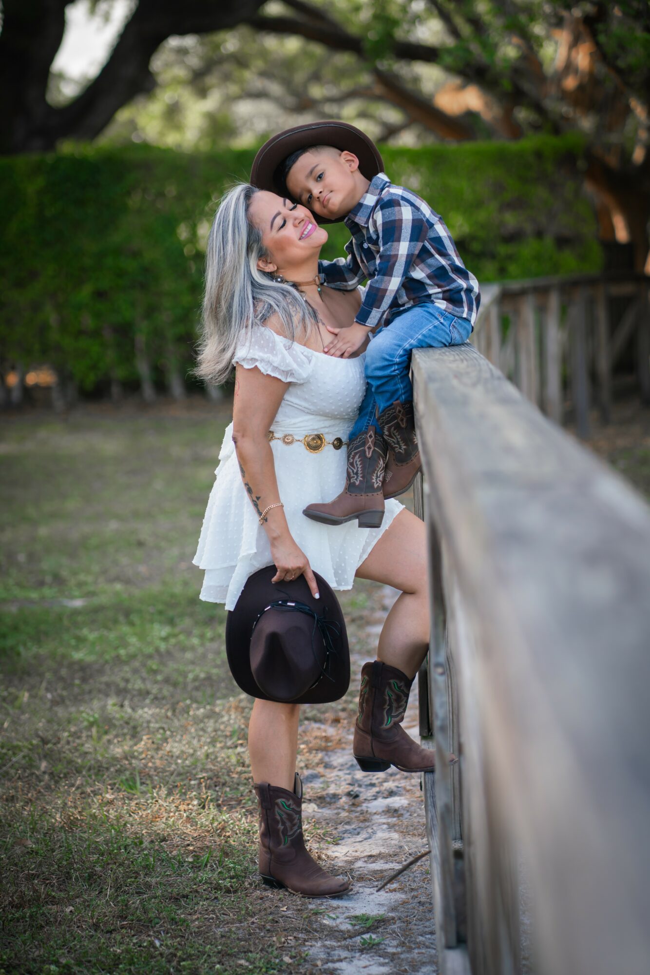 Woman and child leaning on a wooden fence outdoors, woman holding a hat, child resting head on woman's shoulder, trees in background.