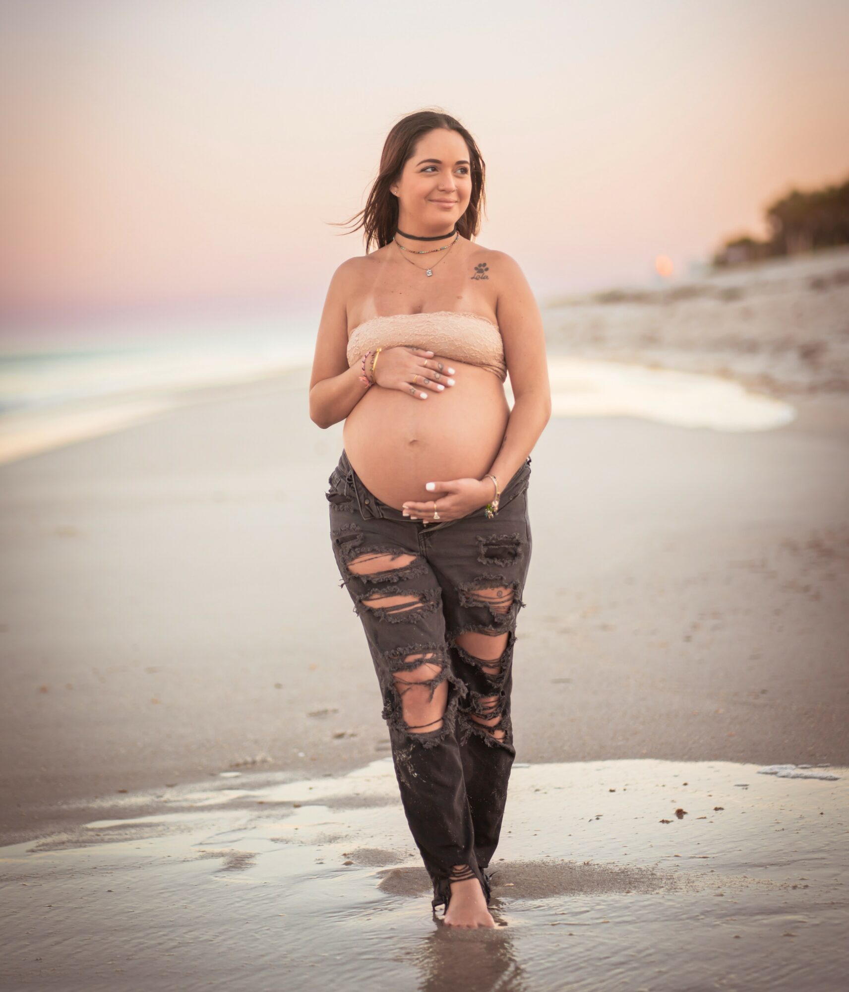 Pregnant woman standing on beach at sunset, smiling, wearing ripped jeans and a strapless top, holding her belly with both hands.