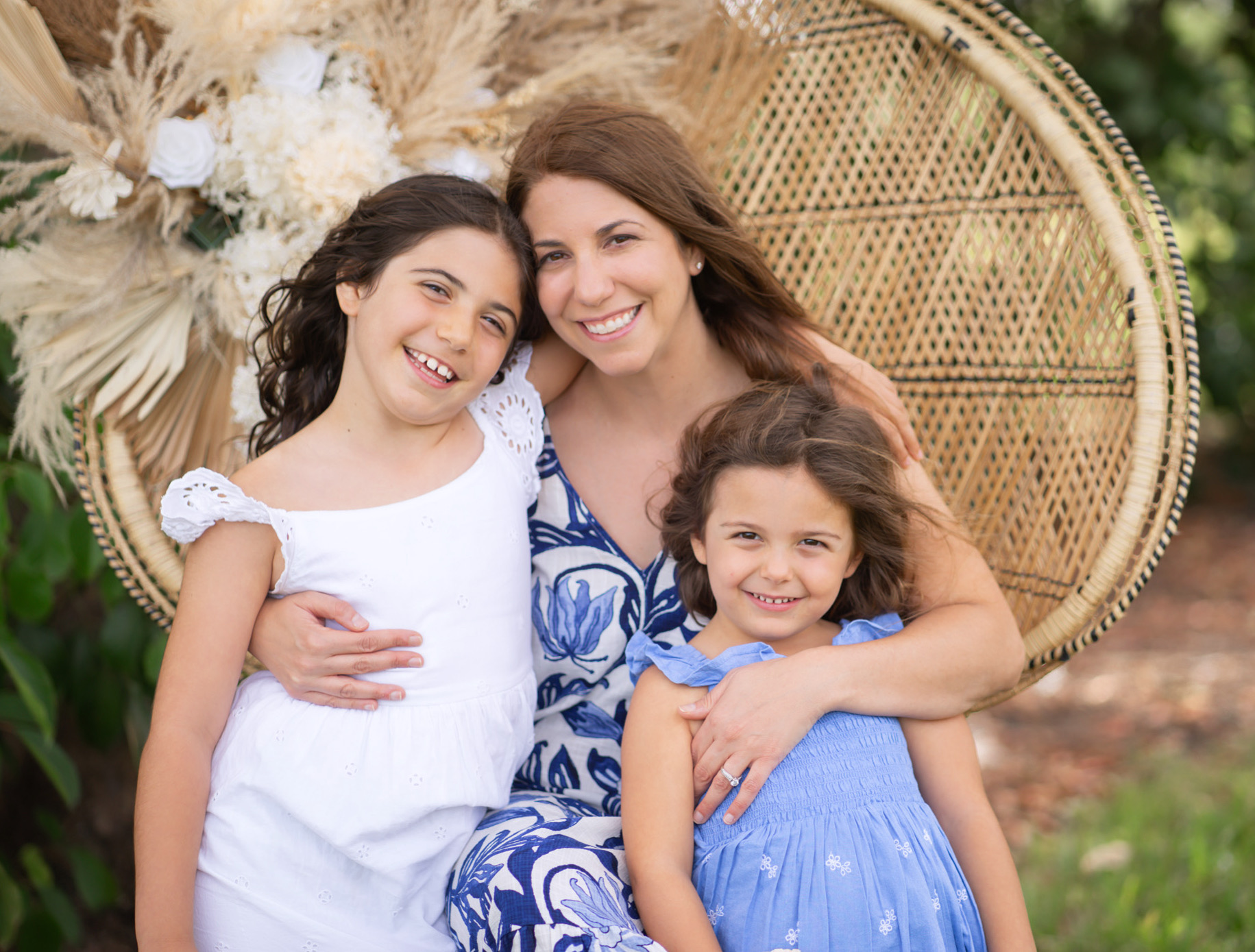 Three smiling females, one adult woman and two young girls, sitting outdoors in front of a woven chair, hugging each other.