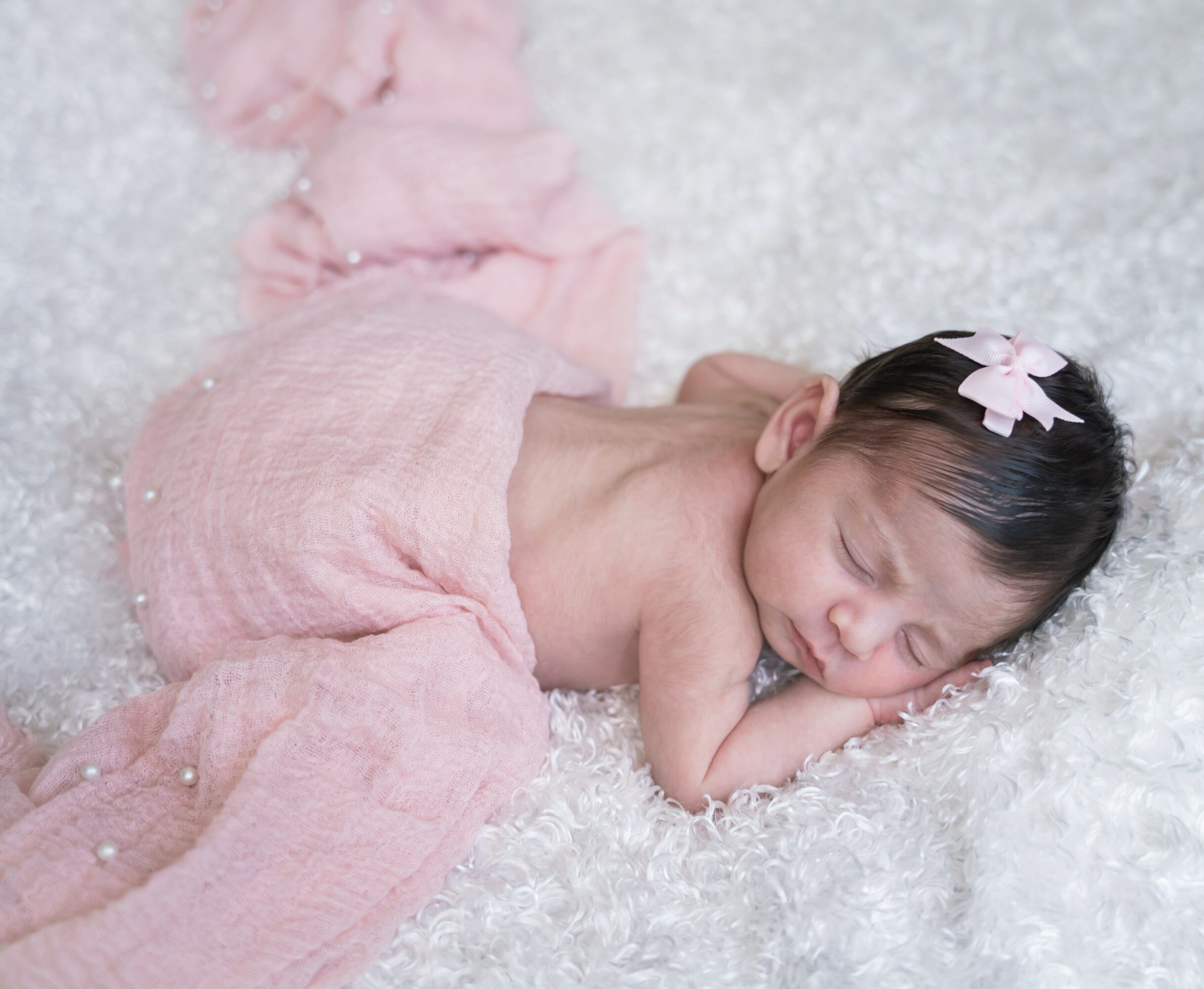 Baby girl lying on her stomach on a soft white surface, wearing pink clothing and a pink bow in her hair, sleeping peacefully.