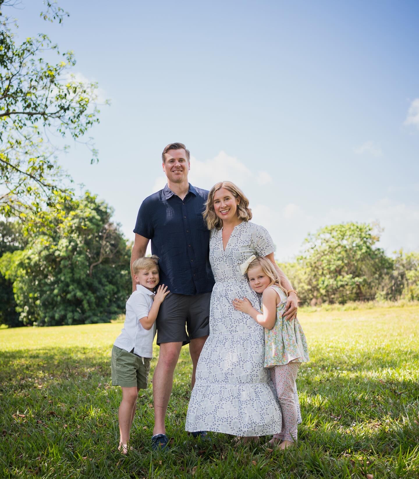 Family of four standing outdoors on grass with trees and blue sky, smiling at the camera.