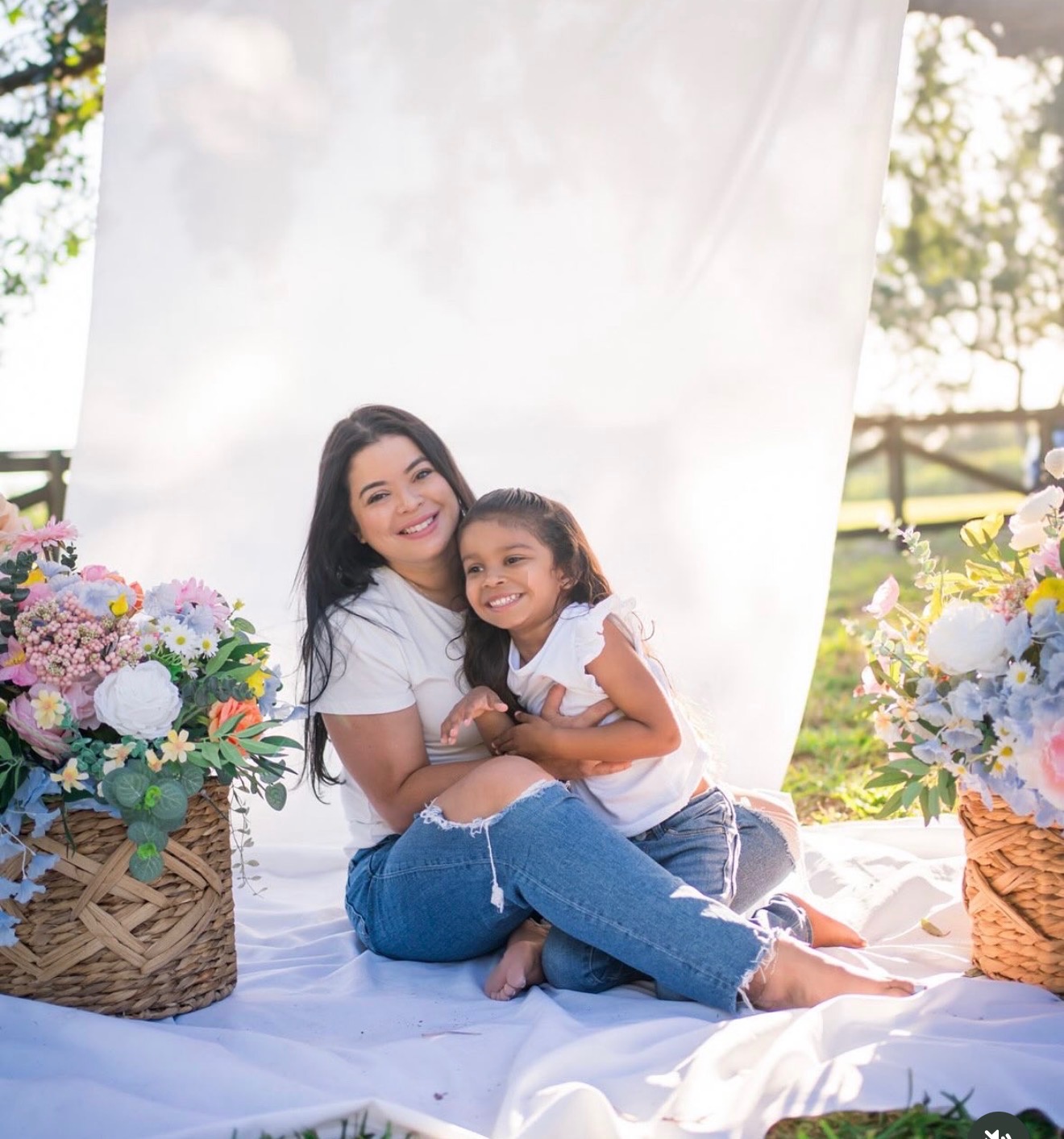 Two smiling females, one adult and one child, sitting on a white blanket outdoors with flower baskets on each side.