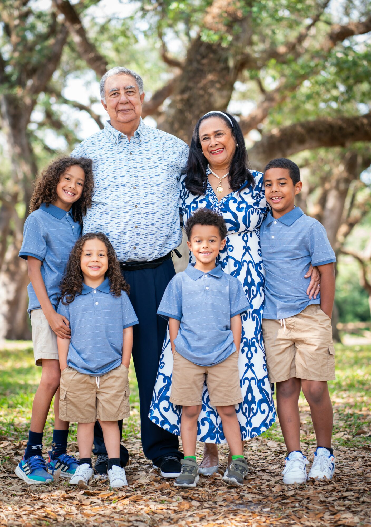 Family of six outdoors with trees, smiling, standing close together, children in blue shirts and khaki shorts, adults in patterned clothing.