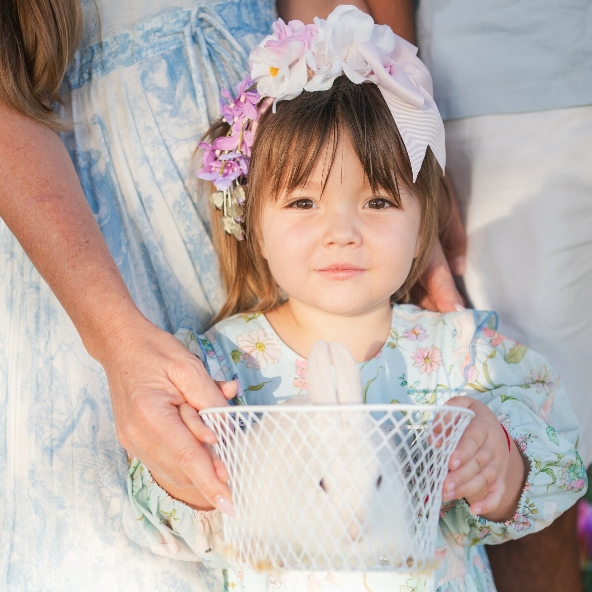 Young girl with a floral headband holding a white rabbit in a wire basket, surrounded by people.