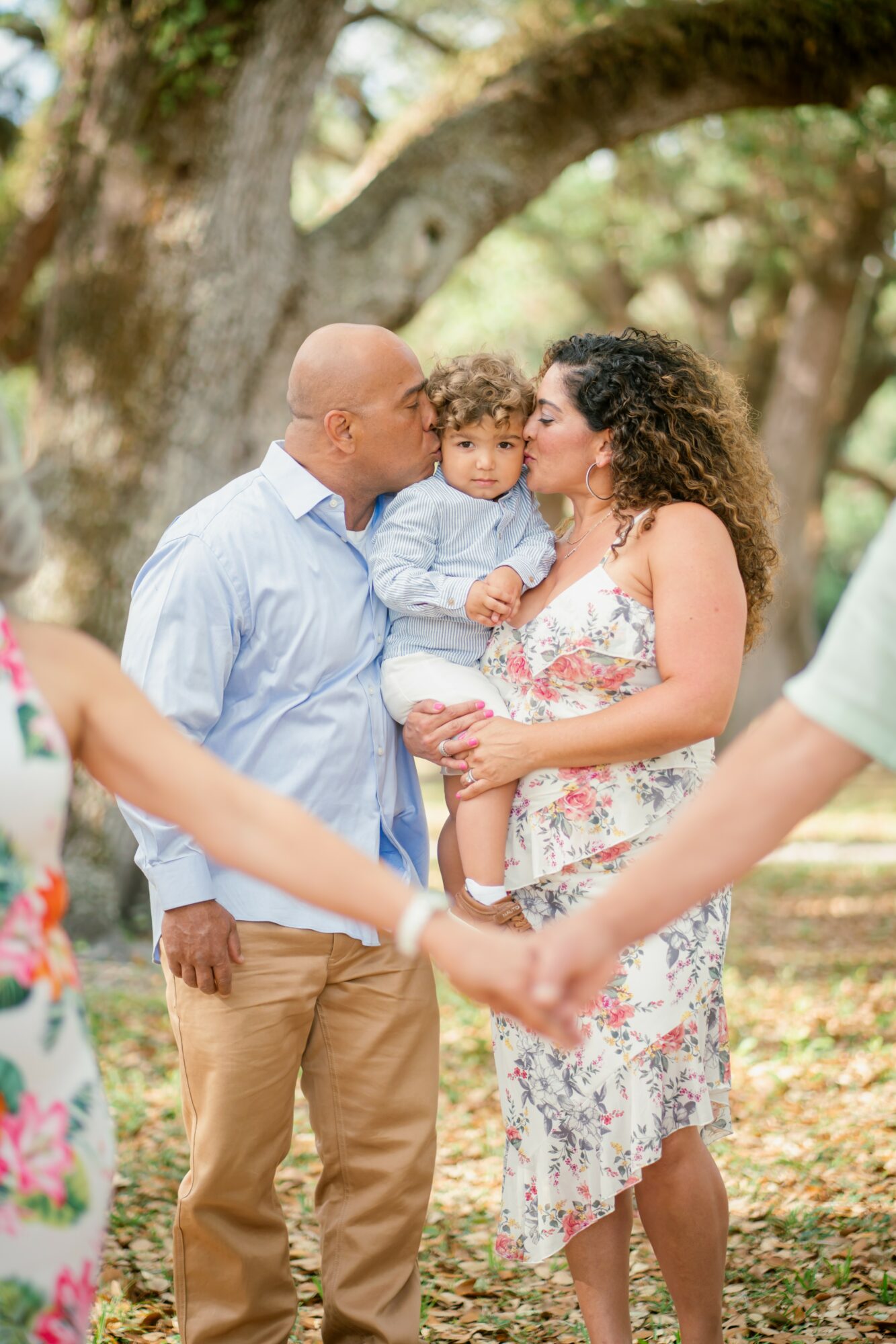 Family outdoors holding hands, with a man, woman, and child near a large tree, smiling and embracing.