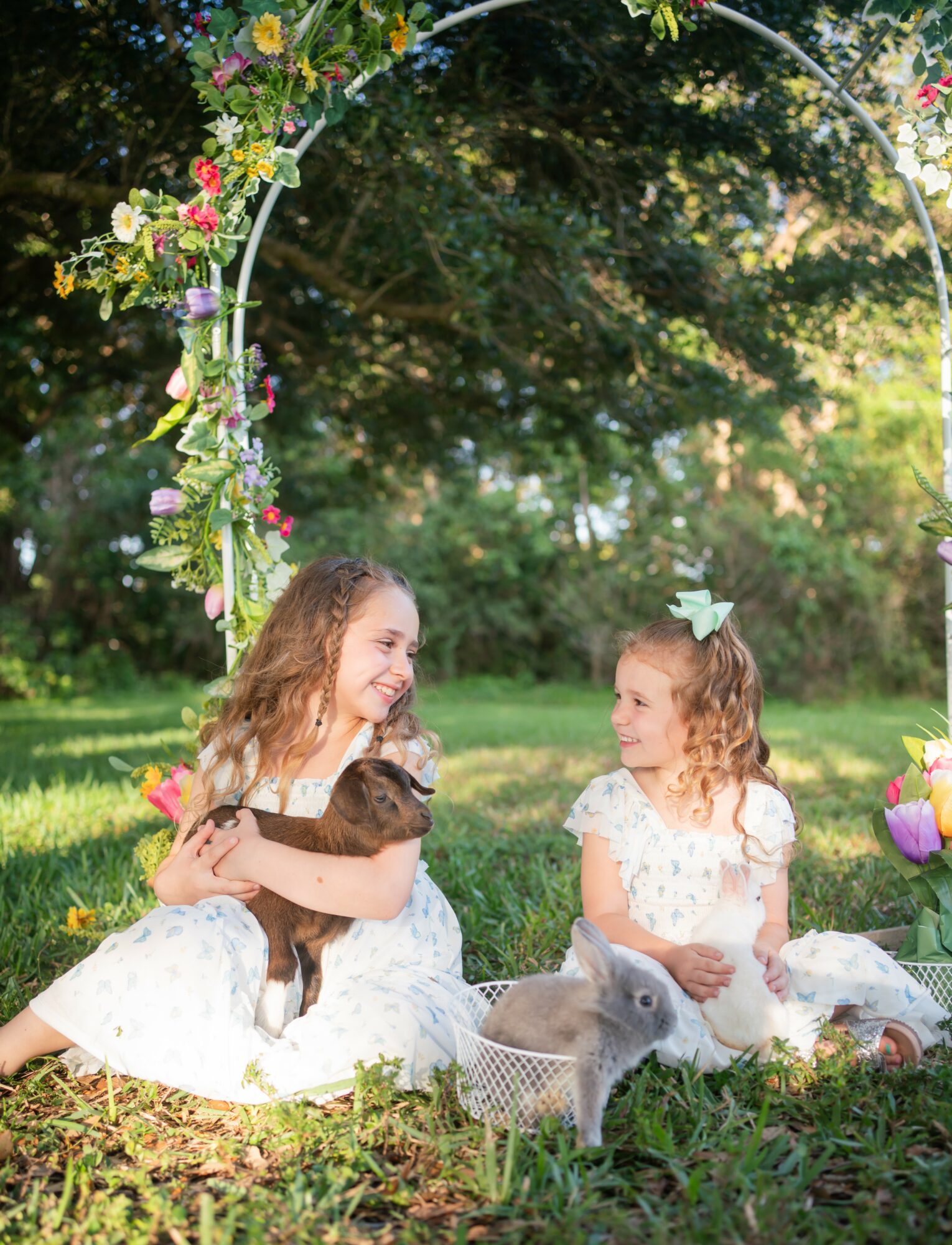 Two young girls sitting outdoors under a floral arch, holding a rabbit, with a duck and a rabbit nearby, in a grassy area.