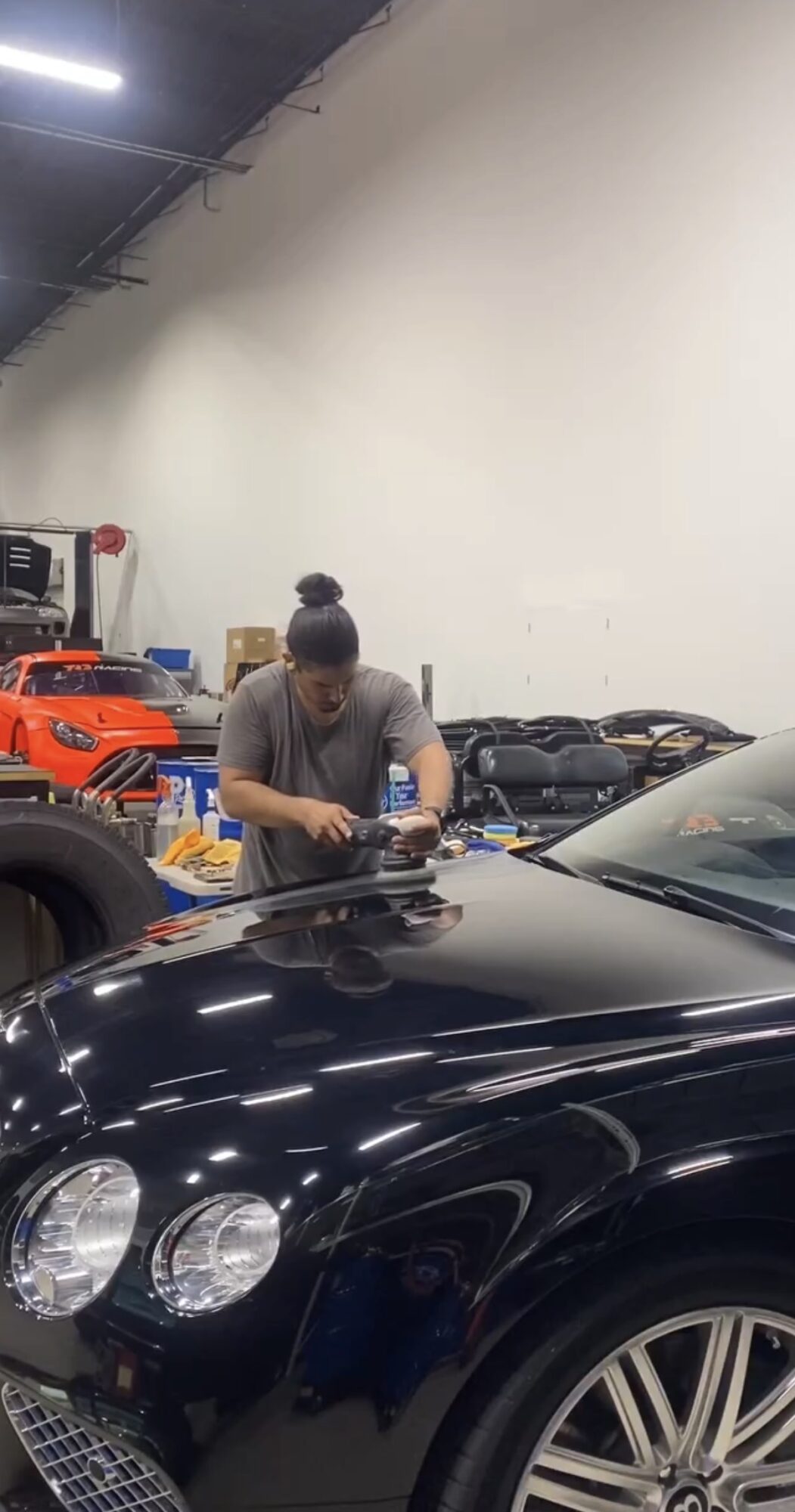 Person with hair in bun working on a car in a garage, surrounded by various vehicles and equipment.