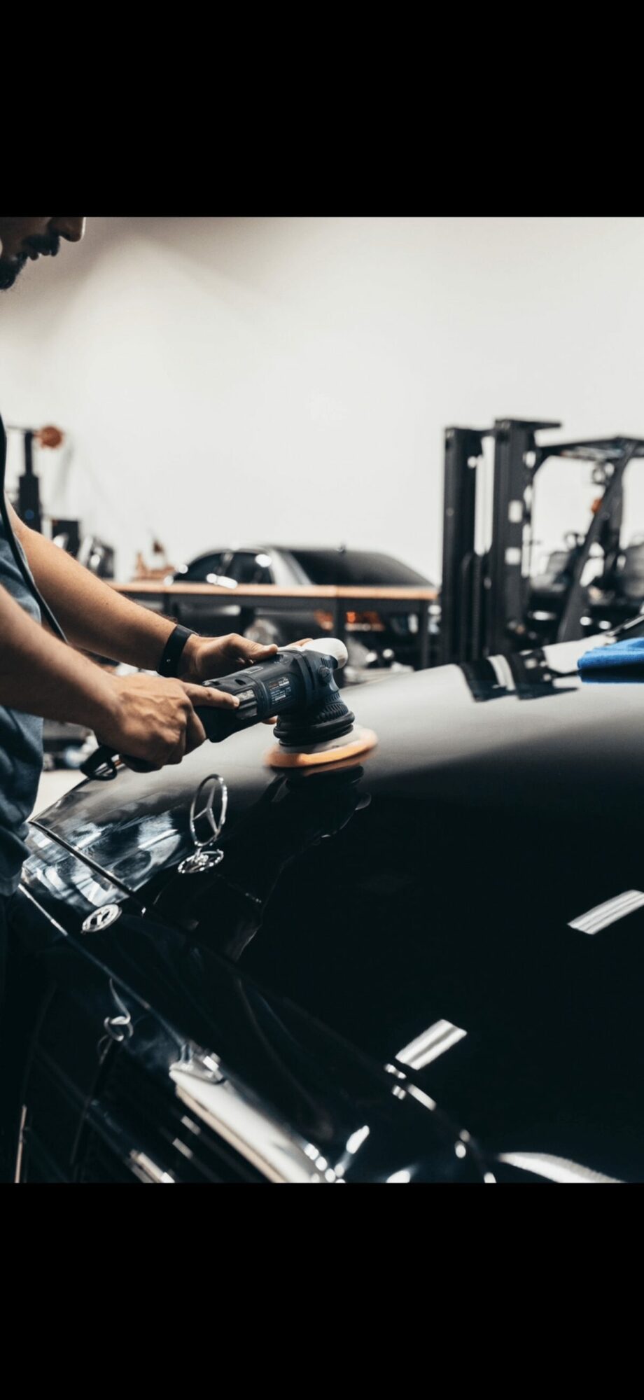Person polishing a black car with a handheld buffer in a garage or workshop setting.
