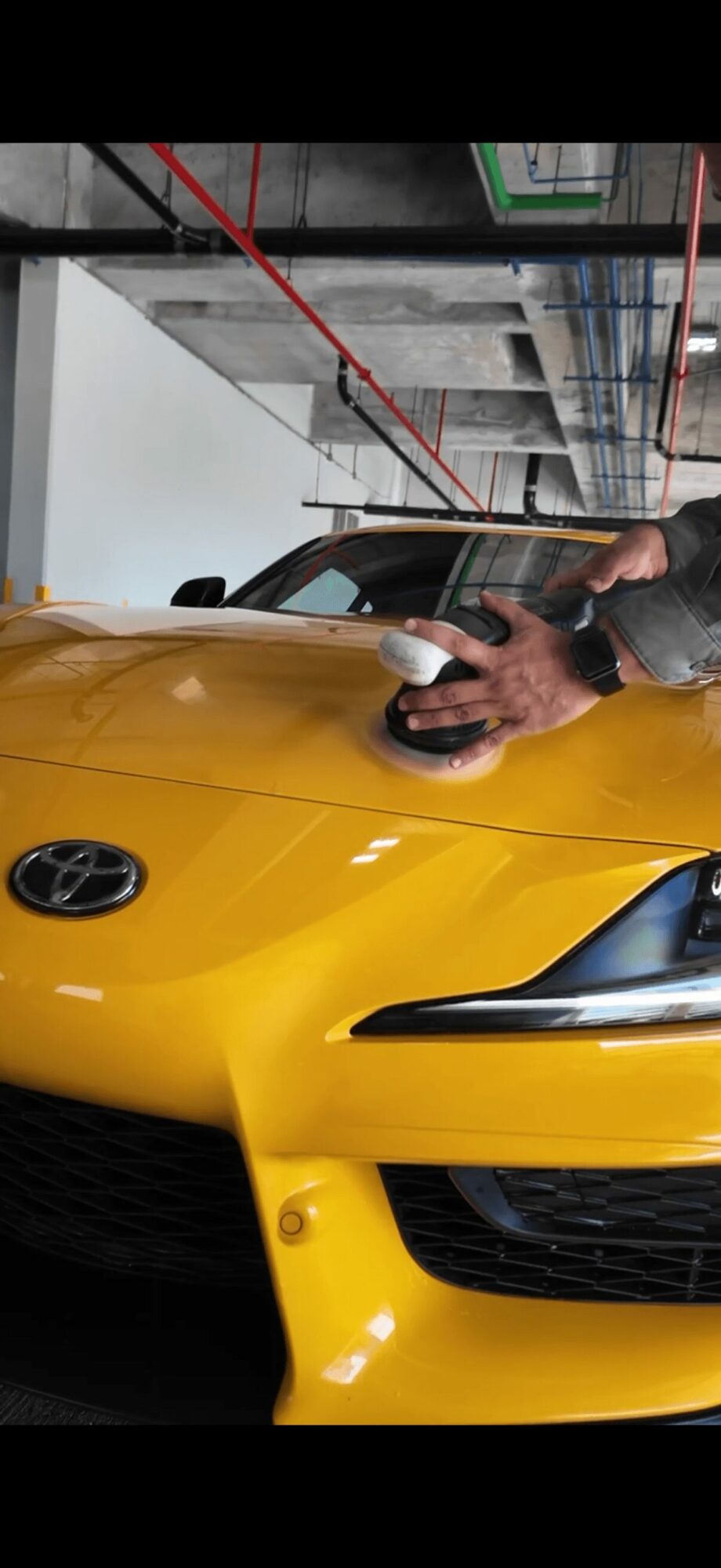 Person cleaning yellow sports car with a cloth, garage setting with ceiling pipes and boxes, hand visible, car's front and side visible.