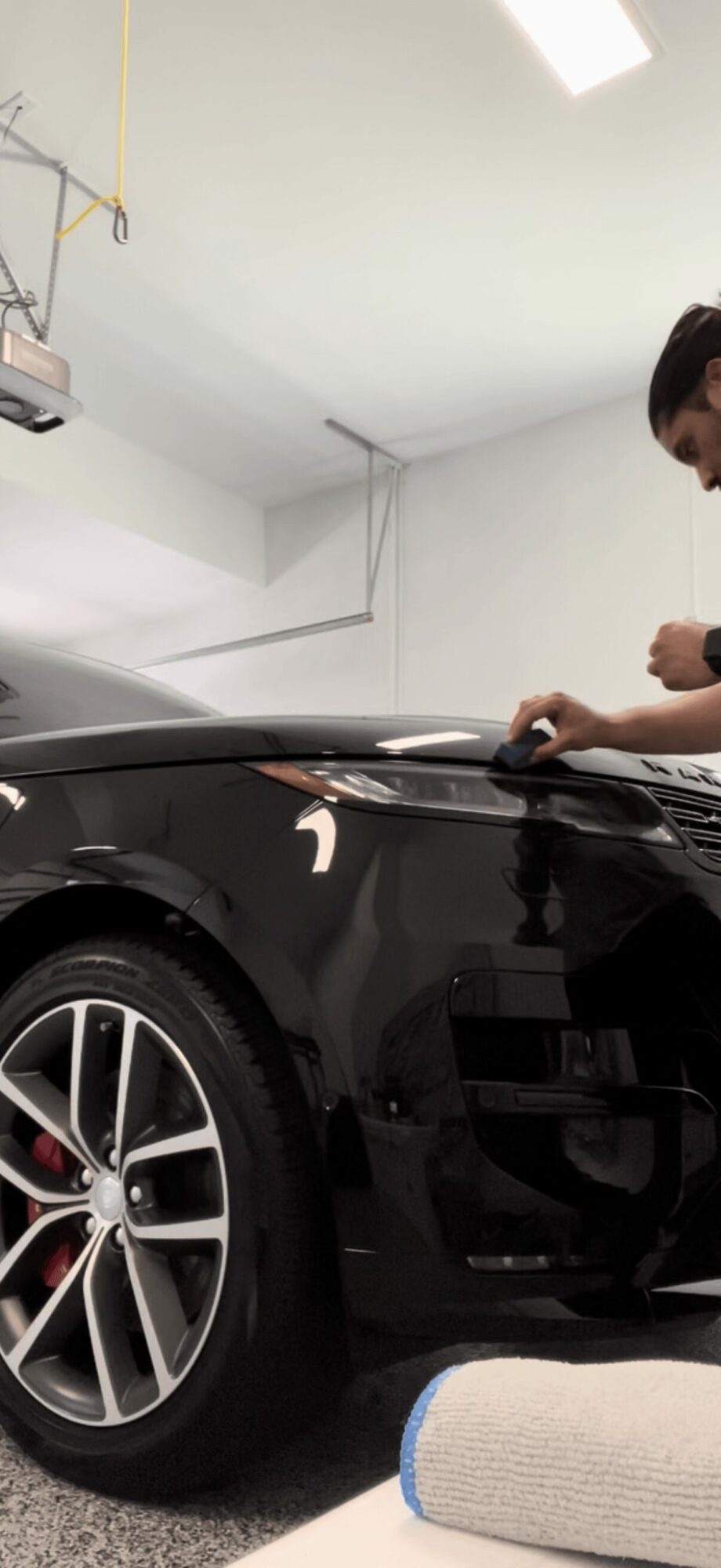 Person working on a black car in a garage, with a white wall and ceiling visible.
