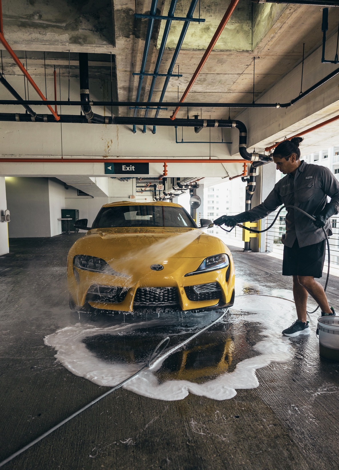 Person washing a yellow sports car in an indoor parking garage with soap and water, reflected on the wet floor.