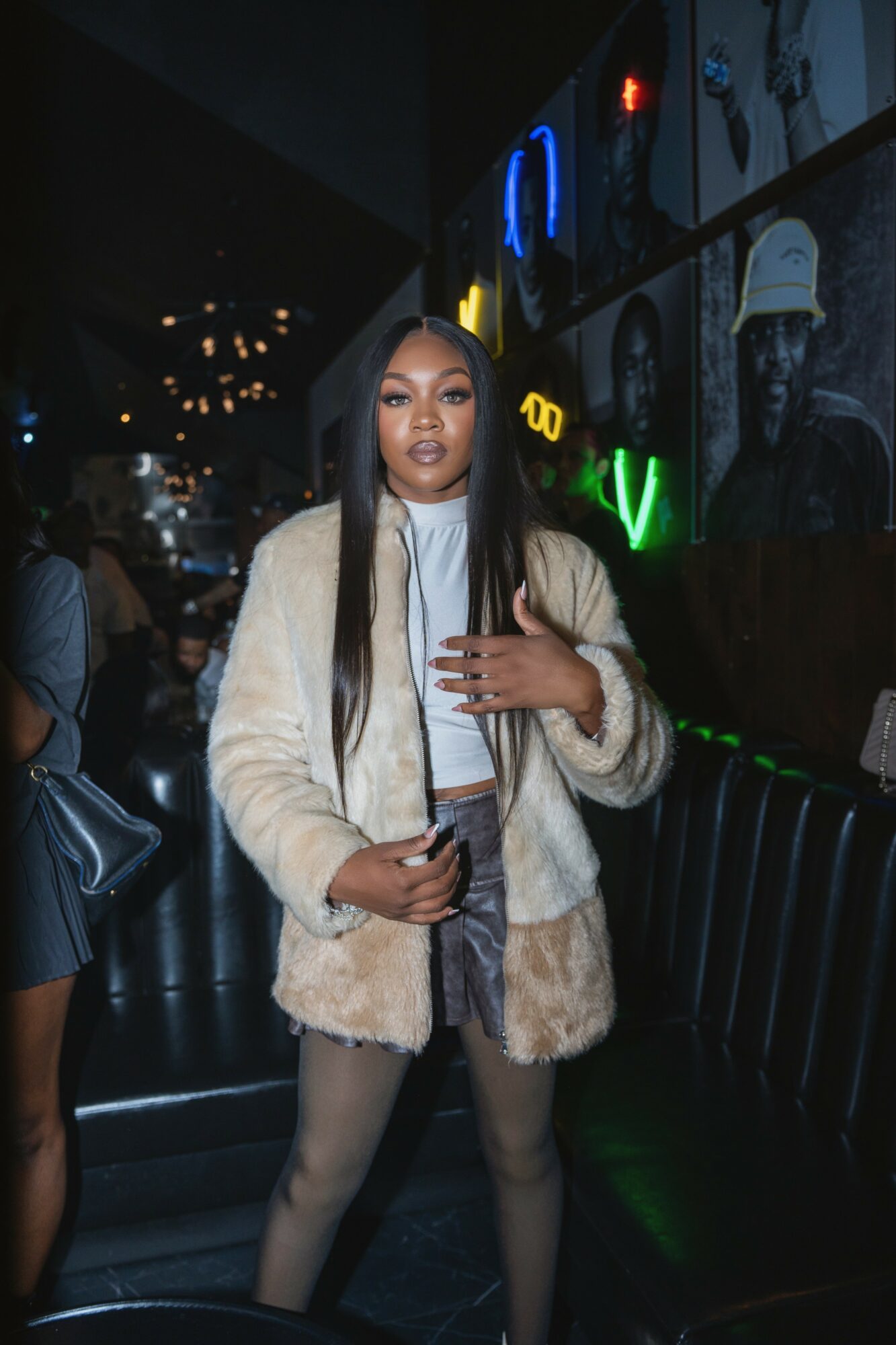 Woman with long dark hair in a beige fur coat, standing in a dimly lit venue with neon signs behind her.