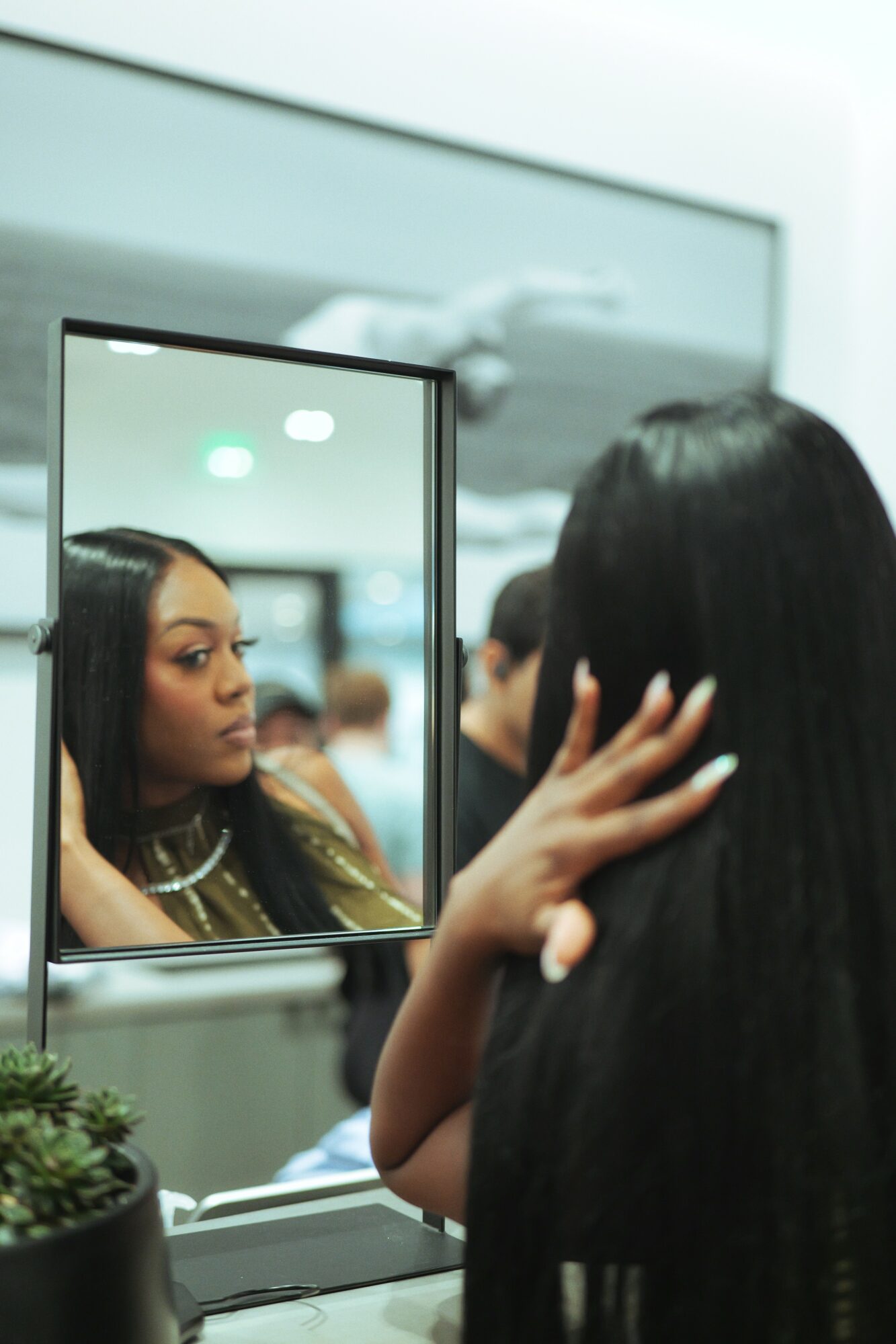 Person with long black hair looking at a mirror, touching their face, with reflection showing their face and makeup, in a salon or similar setting.