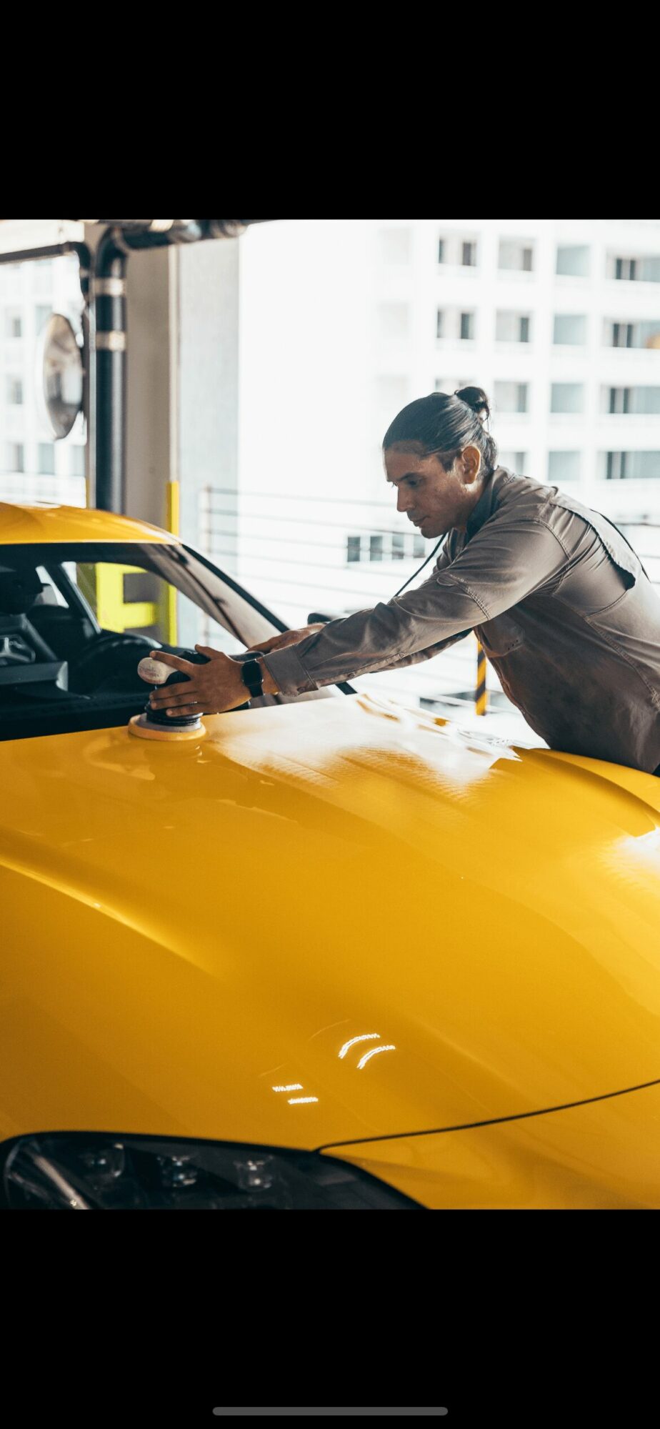 Person cleaning a yellow car inside a building with large windows, cityscape in background.