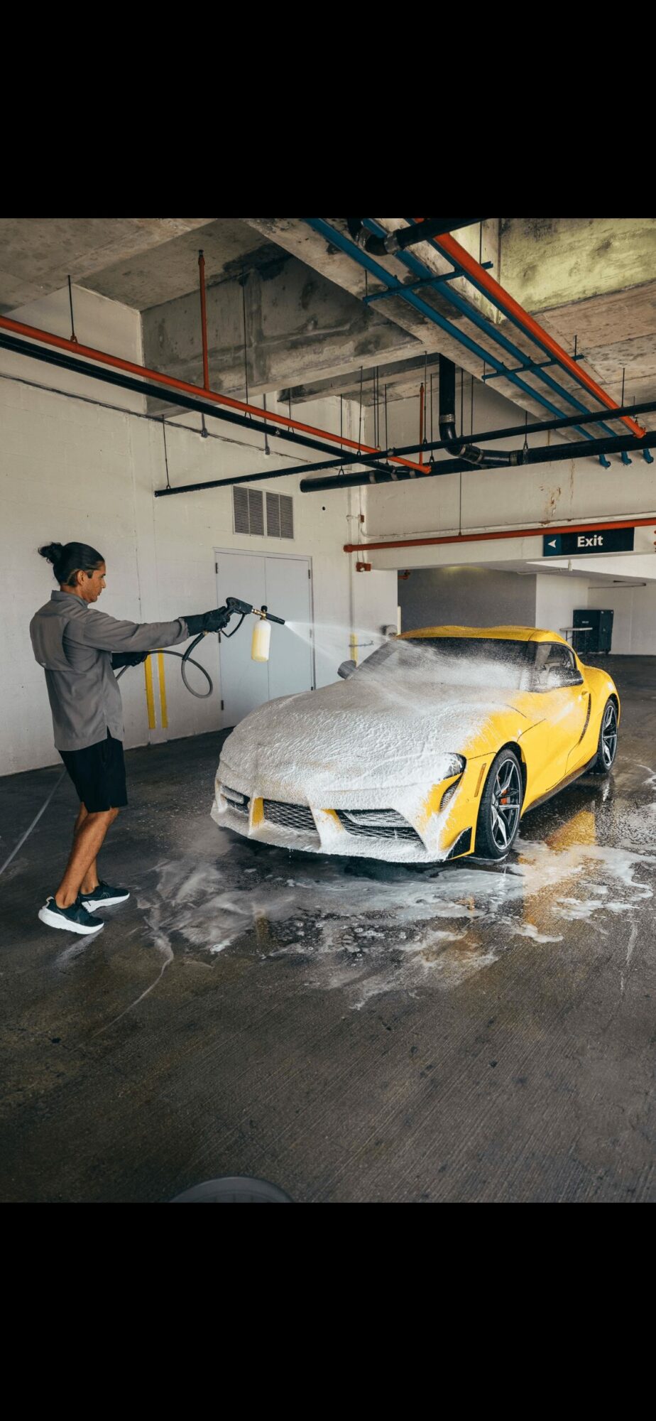 Person washing a yellow sports car with soap in an underground garage, pipes and ceiling visible.