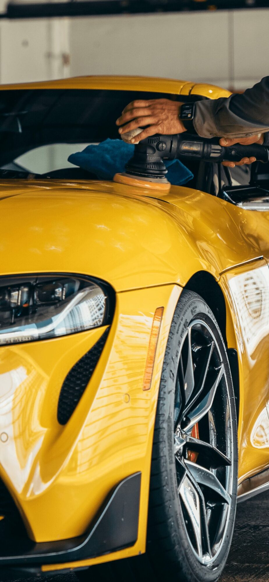Close-up of a yellow sports car with a person working on the engine, hand on the engine bay, black sleeve visible.