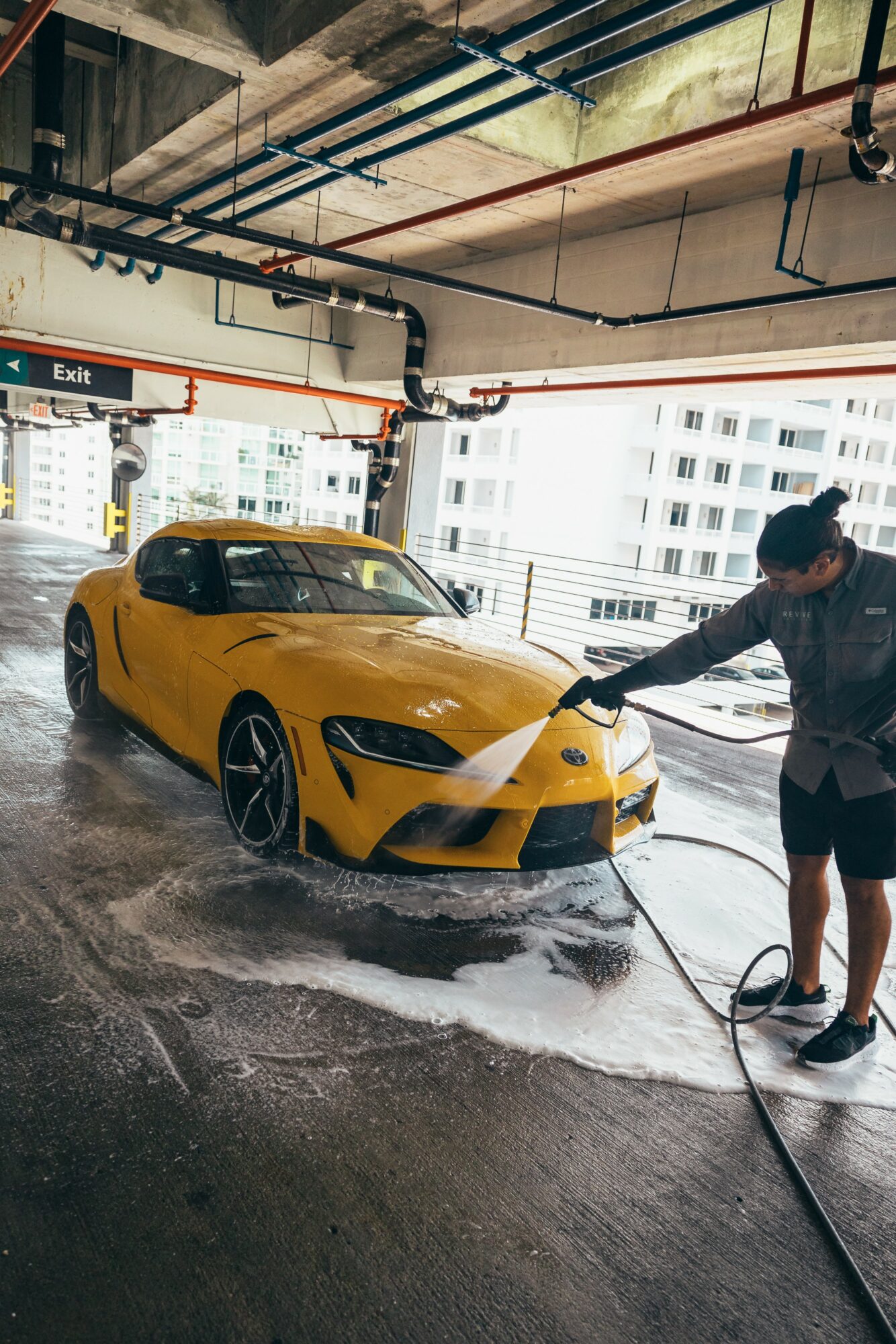Person washing a yellow sports car in a parking garage with city buildings outside, soap and water visible.