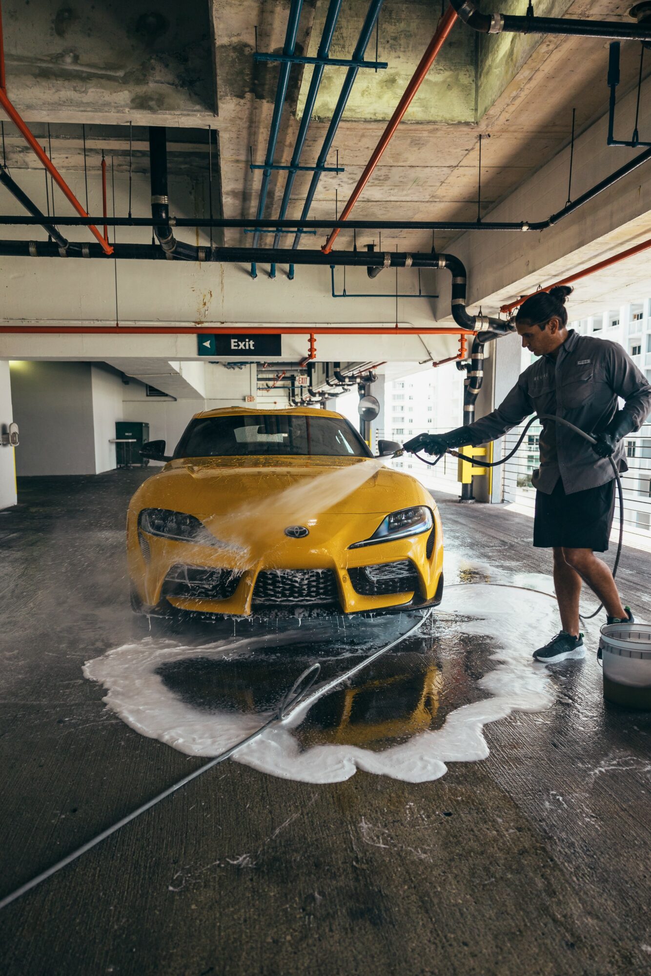 Person washing a yellow sports car in an underground parking garage with water and soap on the floor.