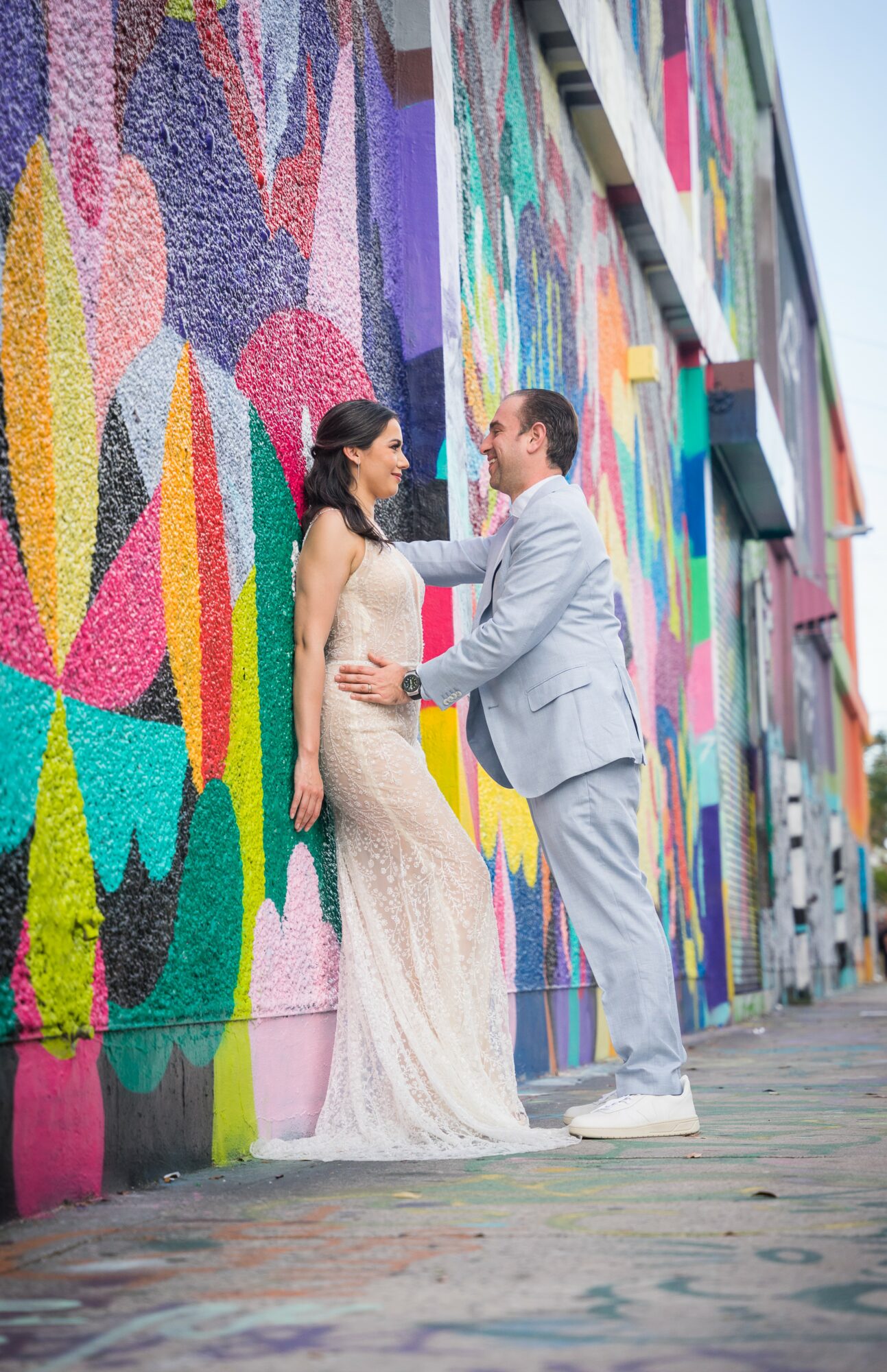 Couple in wedding attire standing close against a colorful mural wall, holding hands and looking at each other.