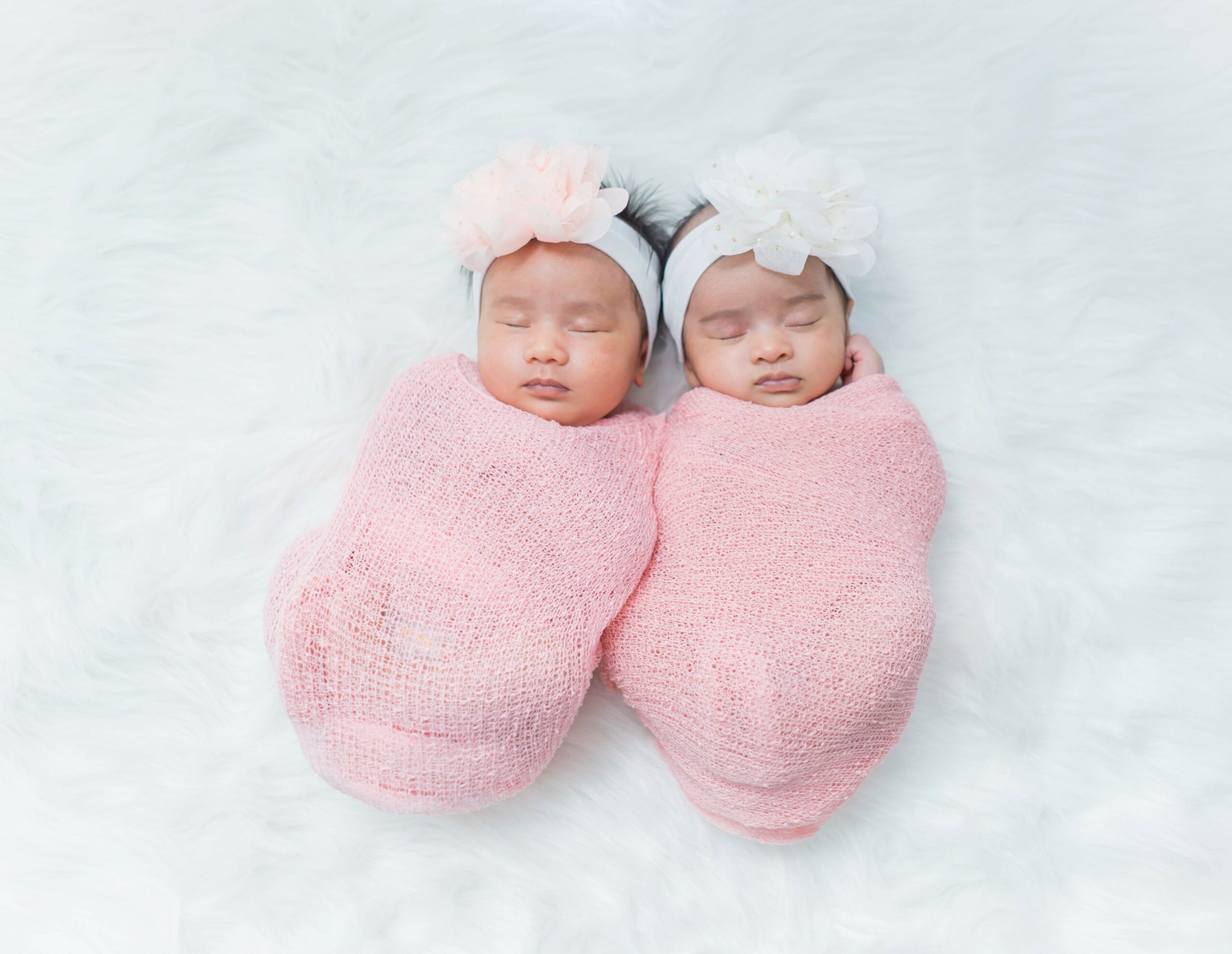 Two newborn babies wrapped in pink blankets, lying side by side on a white surface, wearing headbands with flowers.