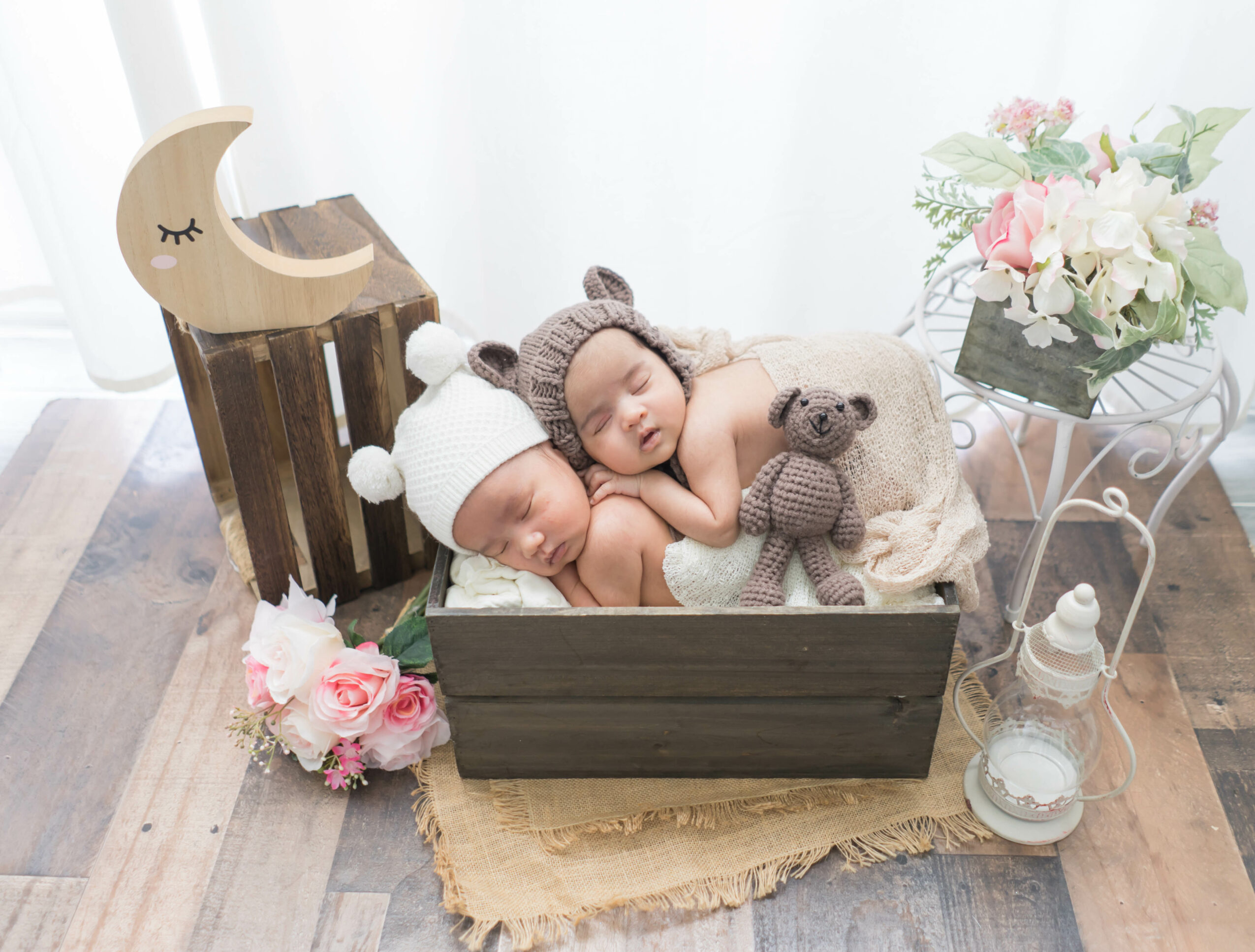 Two sleeping babies in a wooden box, wearing knitted hats, surrounded by flowers and decorative items, on a wooden floor.